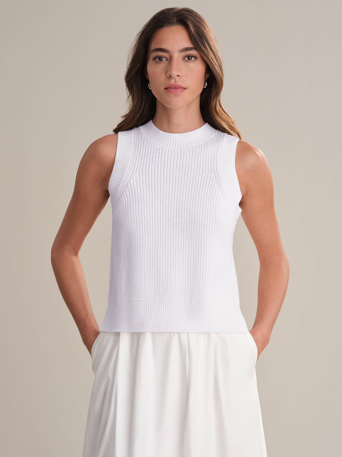 A woman faces forward with hands in her pockets, wearing a 525 America 100% cotton white sleeveless knit top and matching skirt. The crew neck design enhances the versatile look against a plain, light background.