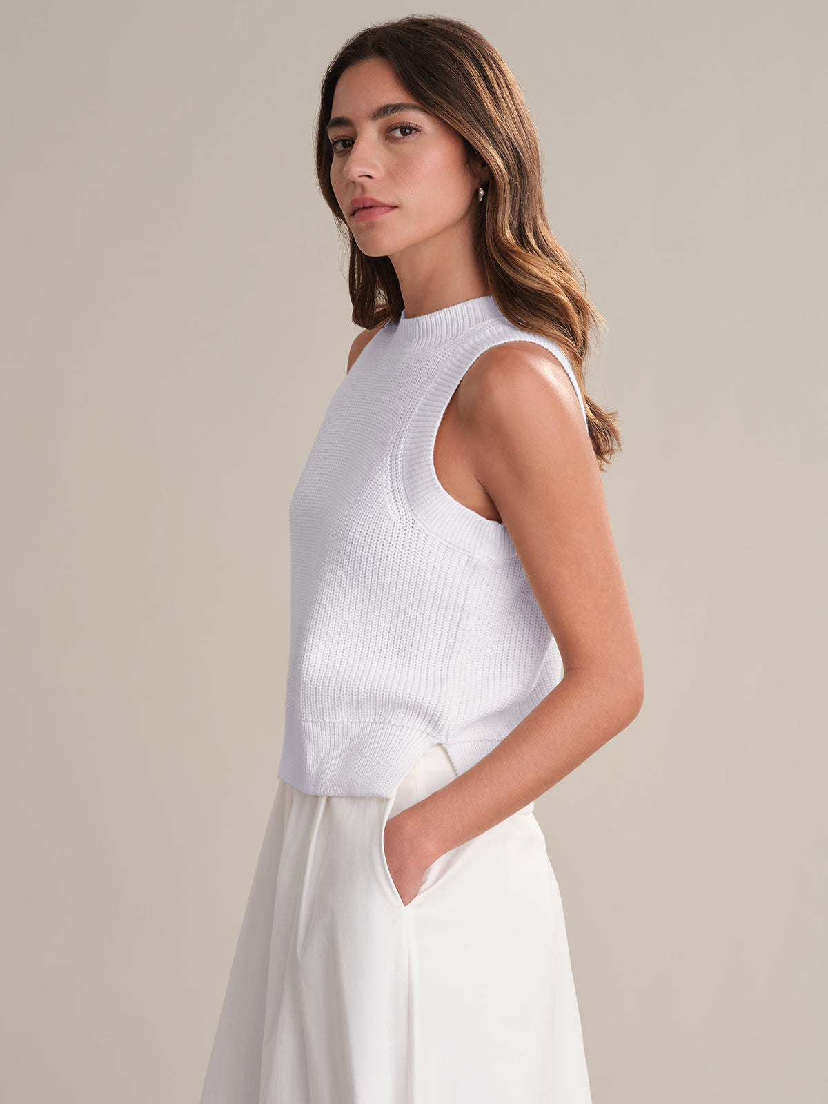 A woman with wavy brown hair stands against a plain backdrop, wearing a 525 America sleeveless white cotton knit crew neck top and matching white pants. One hand is in her pocket as she gazes slightly toward the camera.