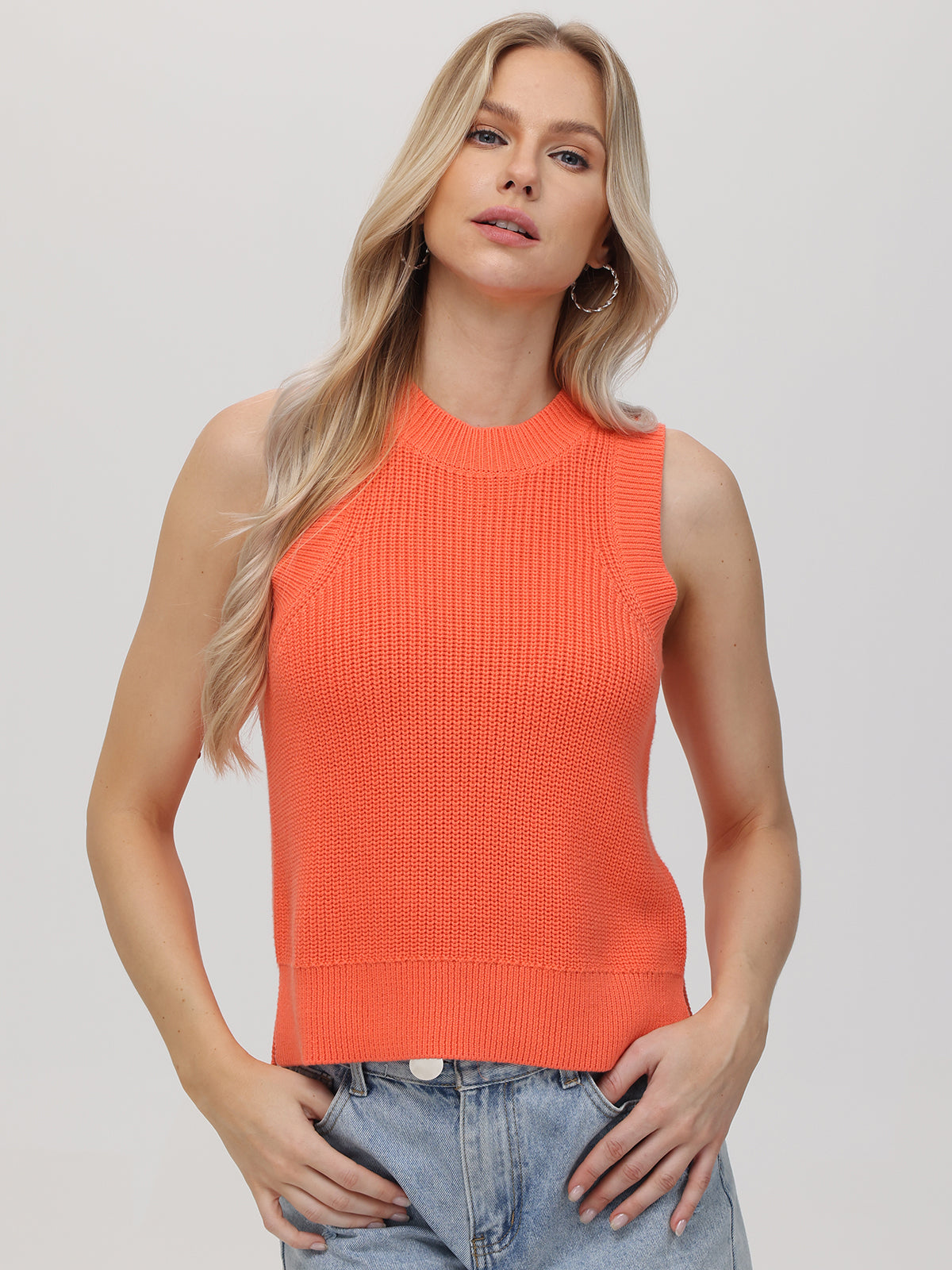 A woman with long blonde hair wears a 525 America sleeveless textured orange knit top with a crew neck and light blue jeans. She stands facing the camera, one hand in her pocket, against a plain light background for a versatile look.