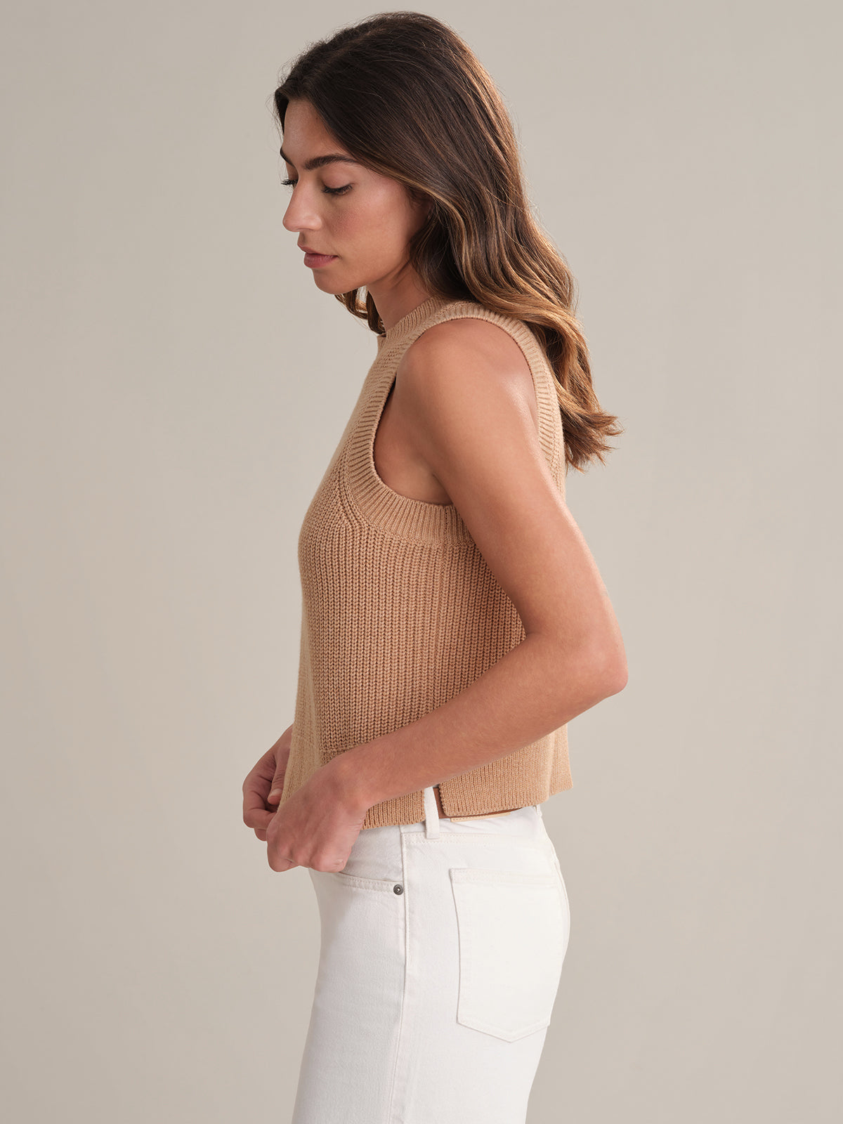 A woman with long brown hair, wearing a 525 America crew neck beige knit top and high-waisted white jeans, stands in profile against a plain light background, embodying casual comfort.