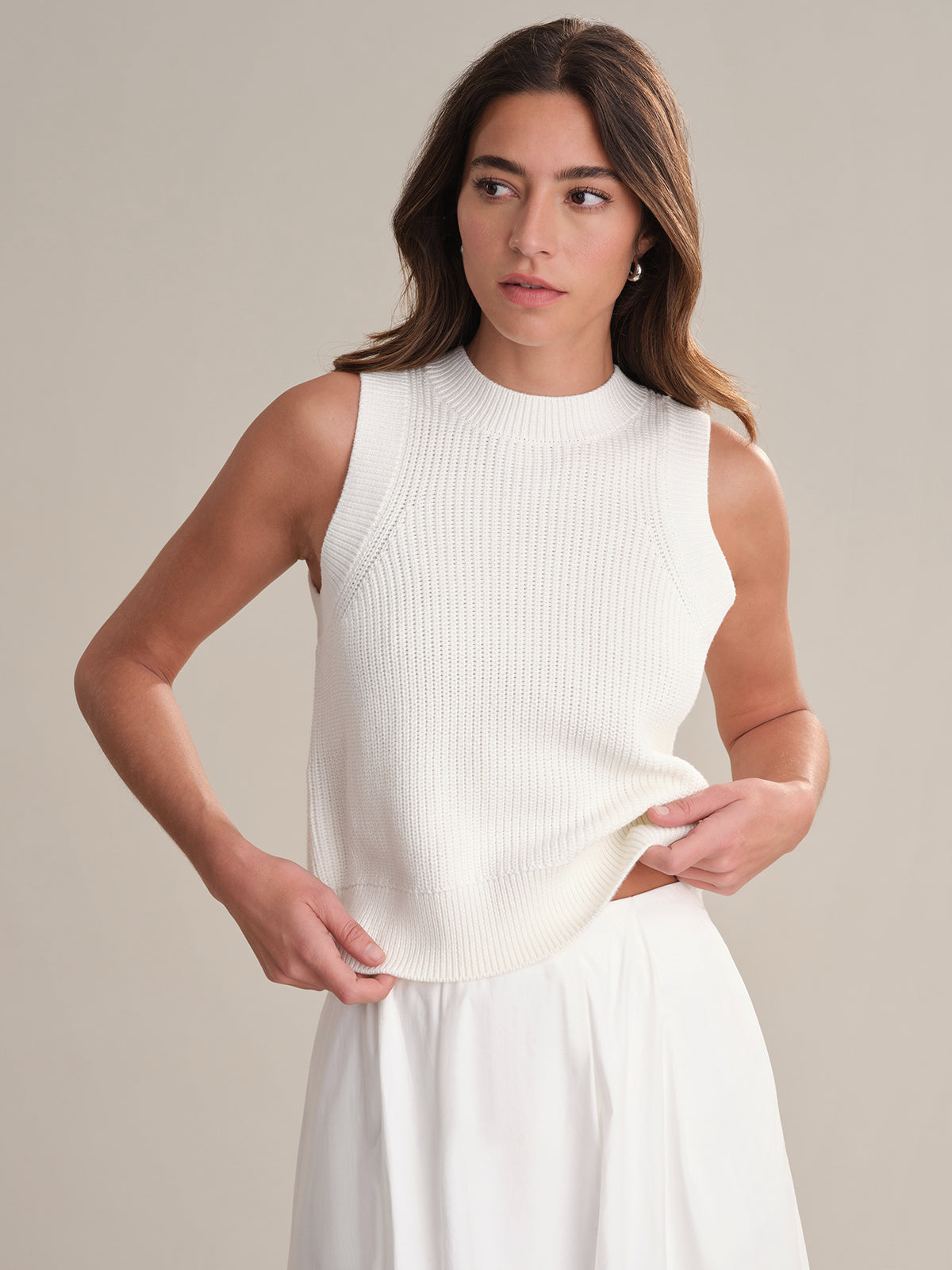 A woman with long brown hair wears a sleeveless white knit crew neck top and a white skirt from 525 America, looking to the side and holding the hem of her top against a plain, light background.
