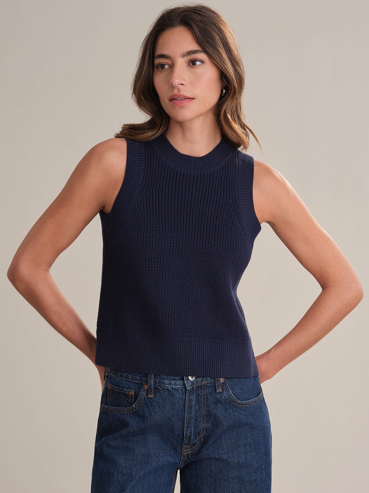A woman models a 525 America sleeveless navy blue 100% cotton knit top with blue jeans, standing against a plain light background with her hands behind her back, highlighting the outfits versatile style.