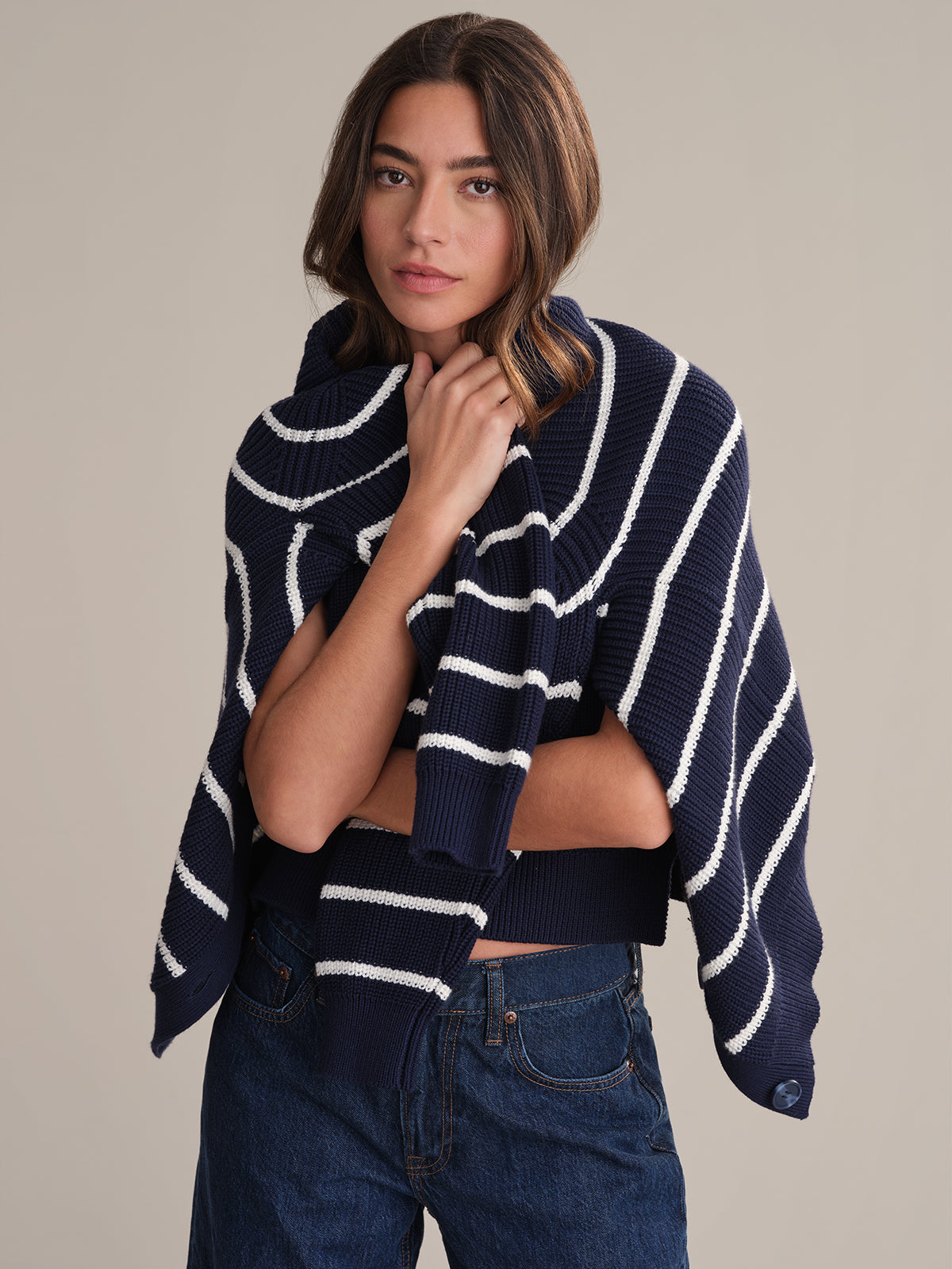 A woman with shoulder-length brown hair wears a 525 America relaxed fit navy blue sweater with white stripes draped over her shoulders, paired with blue jeans. She stands against a plain beige background, looking at the camera neutrally.