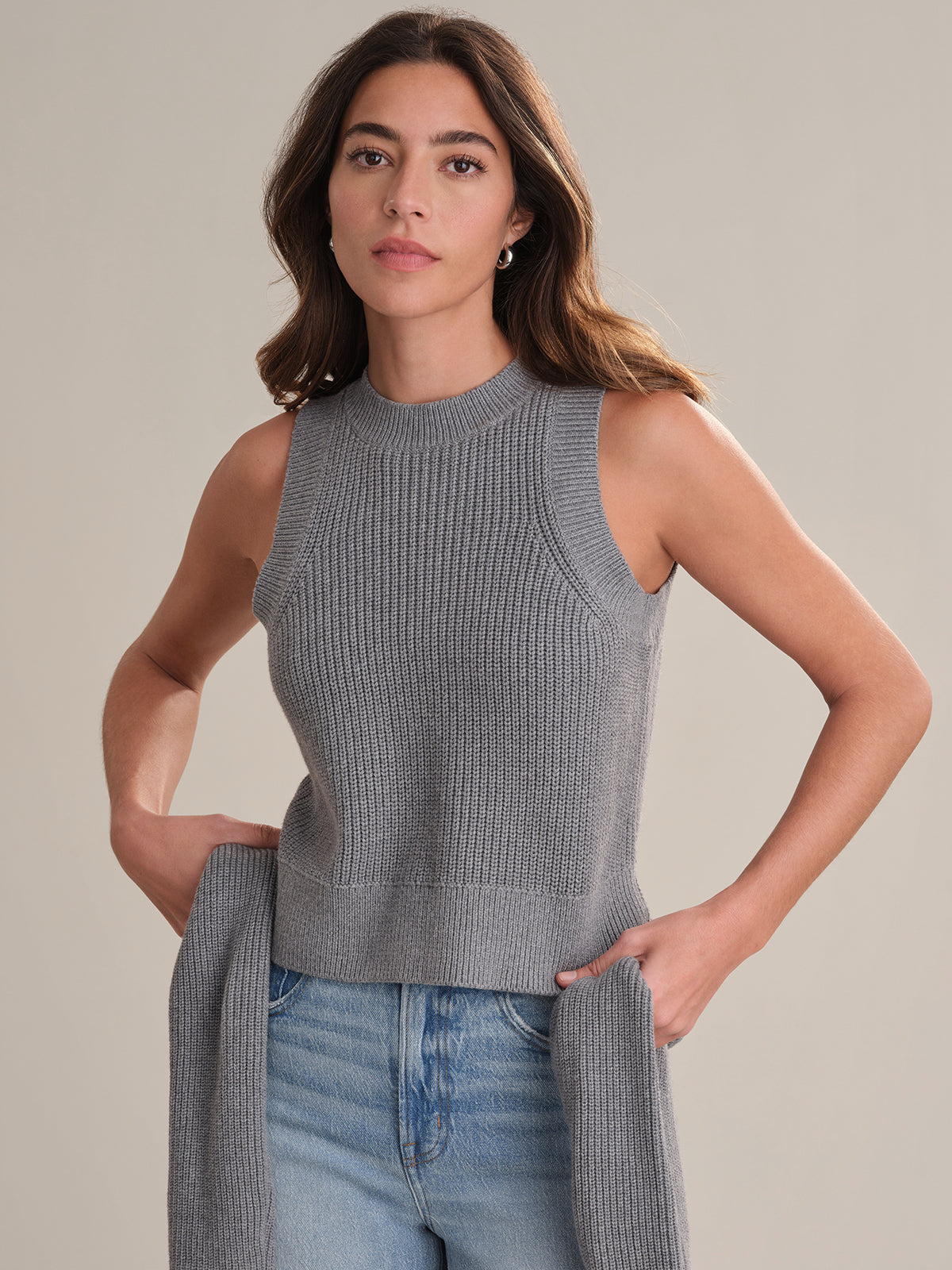 A woman with long brown hair models a sleeveless gray knit crew neck top from 525 America, paired with light blue jeans. She stands against a beige background, holding a matching gray knit piece for added versatility.
