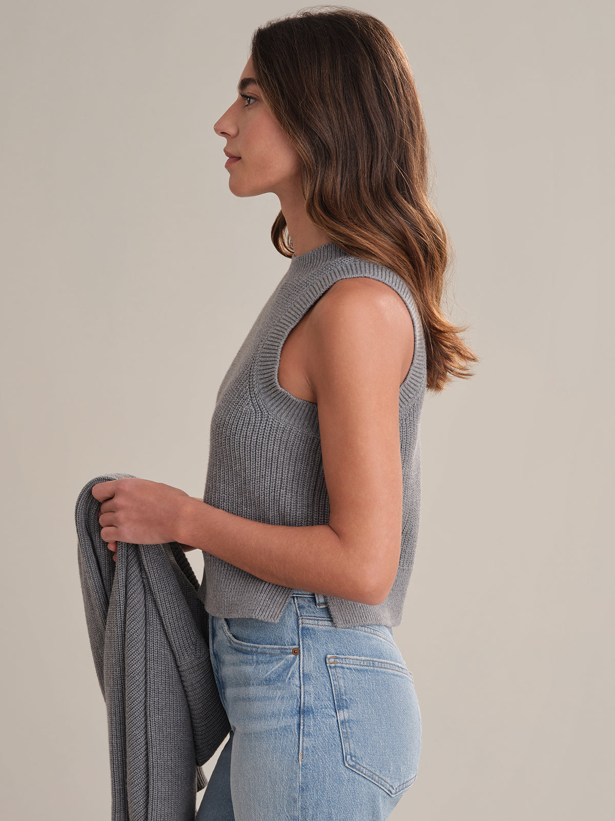 A woman with wavy brown hair stands in profile against a plain background, wearing a 525 America gray sleeveless crew neck knit top and light blue jeans, holding a matching gray garment in her left hand for a coordinated look.