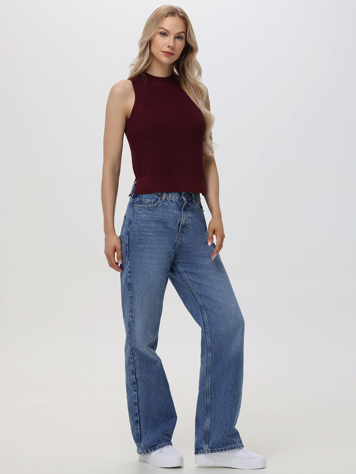 A woman with long, wavy blonde hair stands against a plain light background. She wears a 525 America 100% cotton burgundy sleeveless knit crew neck top, high-waisted wide-leg blue jeans, and white sneakers for a casual look.