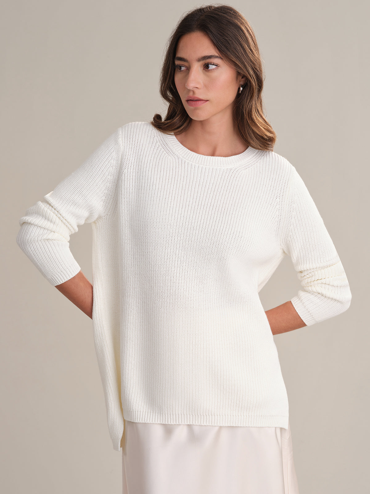 A woman models the 525 America relaxed fit, long-sleeved white shaker stitch knit sweater, posing against a plain background with her hands on her hips and gazing to the side.