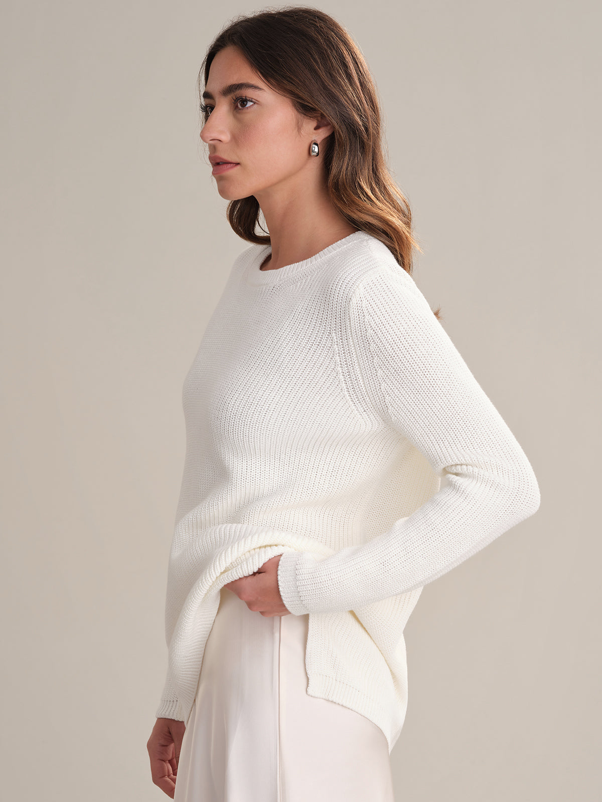 A woman with long brown hair stands in profile against a plain background, wearing a white 100% cotton shaker stitch relaxed fit sweater by 525 America and light-colored pants. She looks ahead neutrally, holding the sweaters hem.