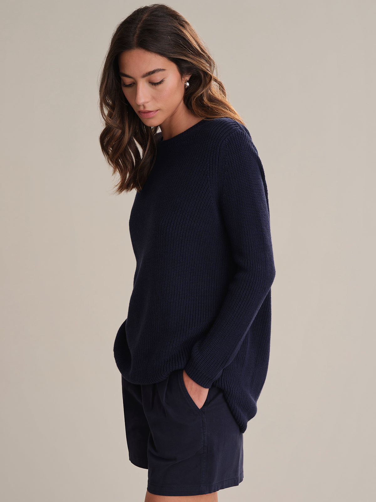 A woman with wavy brown hair looks down while wearing a 525 America relaxed fit, dark navy shaker stitch knit sweater and matching navy shorts. She stands against a light background, her left hand casually in her pocket.