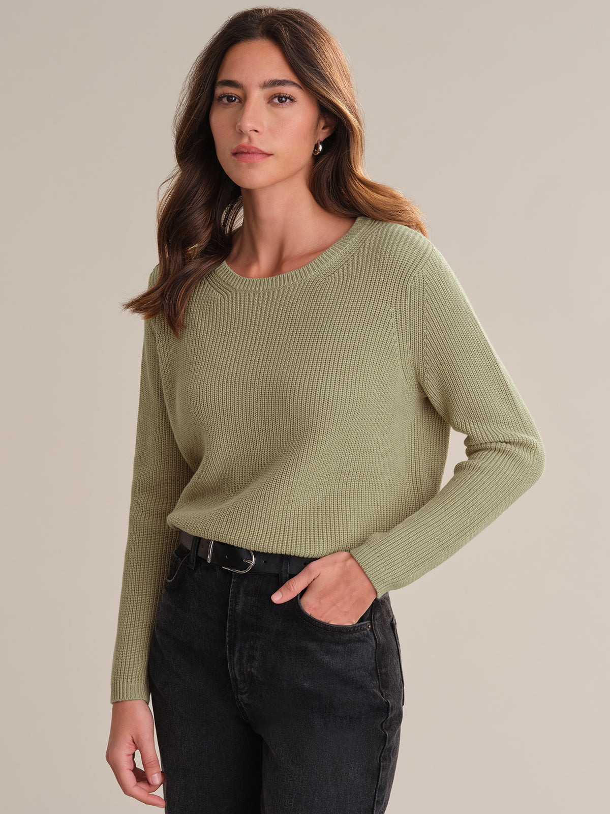 A person with long brown hair wears a 525 America light green shaker stitch sweater with a relaxed fit and high-waisted black jeans, standing with one hand in their pocket against a plain, neutral background.