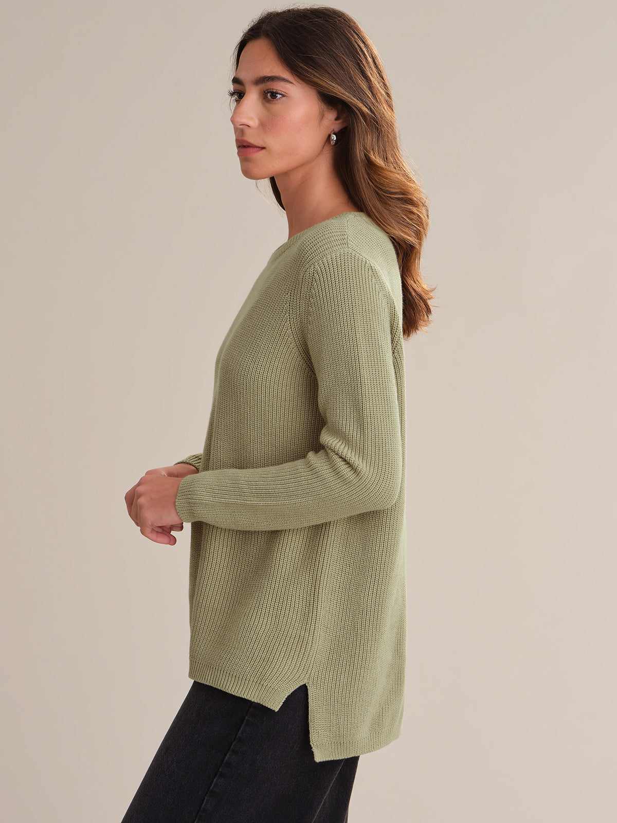 A person with long brown hair wears a 525 America light green shaker stitch sweater in a relaxed fit and black pants, standing in profile against a neutral background.