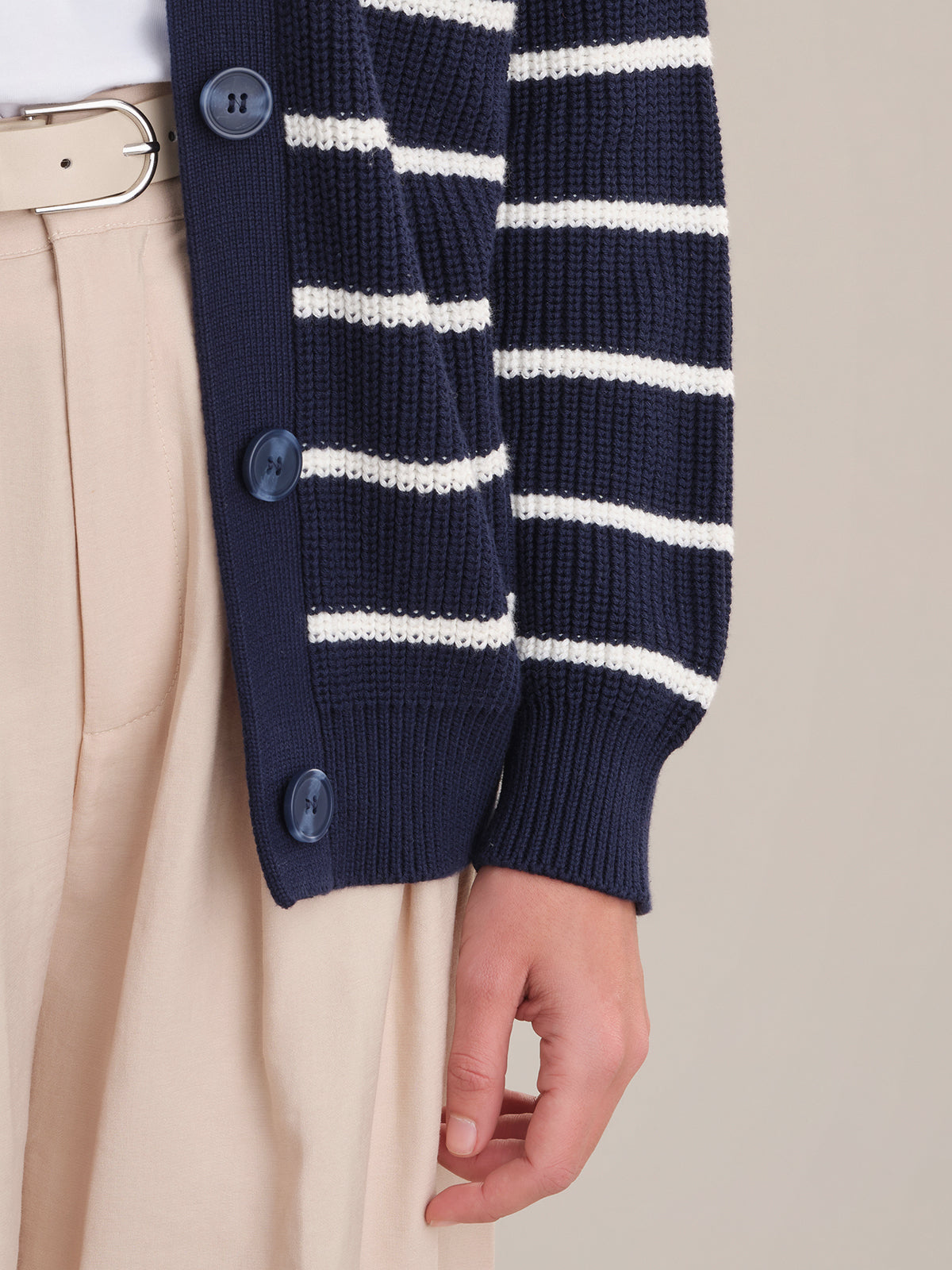 A person wears relaxed fit beige trousers, a light belt, and a 525 America navy blue cardigan with white stripes and blue buttons. Only the lower torso and one hand are visible against a plain background.