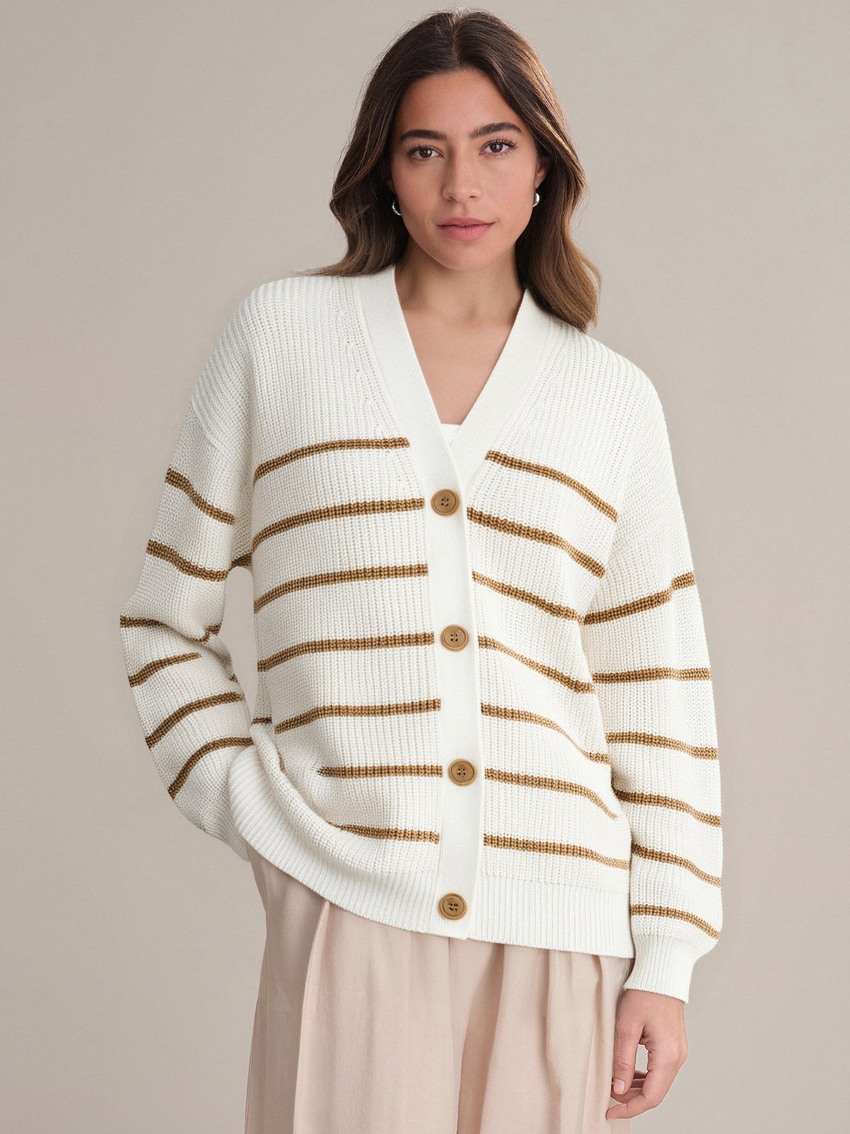 A person wears a 525 America relaxed-fit, white cotton cardigan with horizontal brown stripes and brown buttons, paired with light beige pants, standing against a plain, light background.