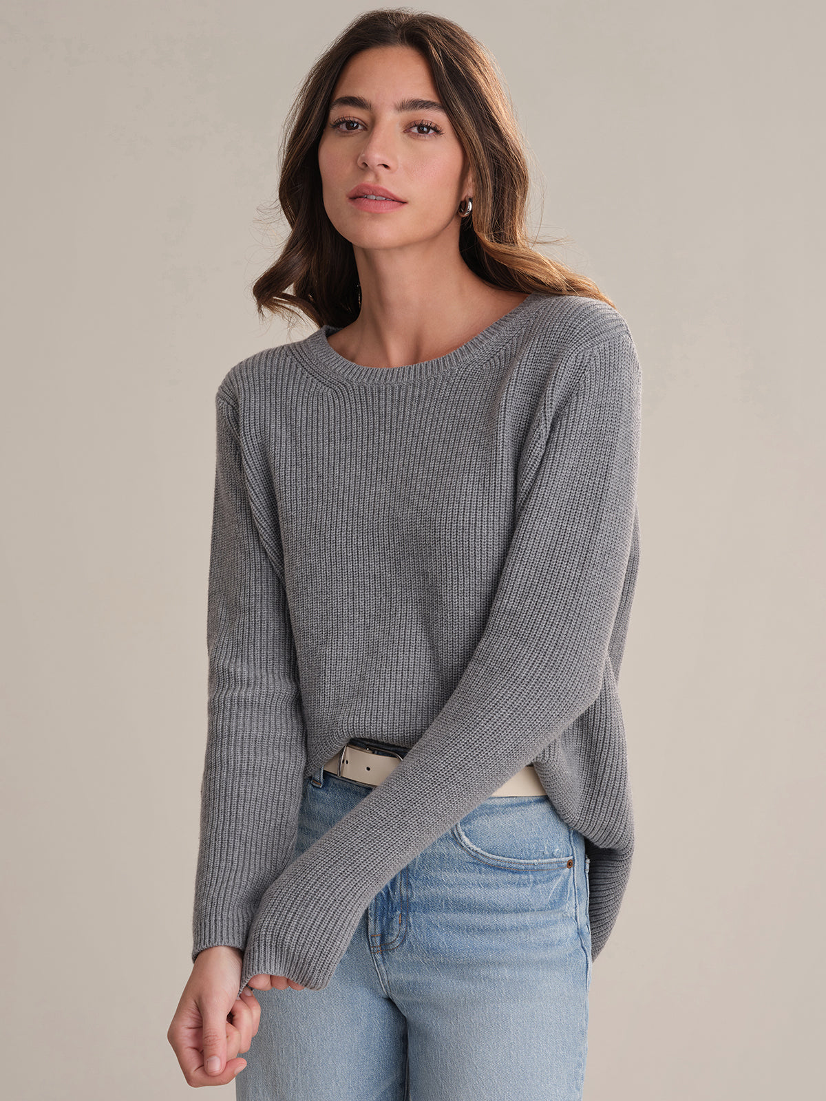 A person with long brown hair wears a gray, 100% cotton ribbed sweater by 525 America with a relaxed fit, light blue jeans, and a light belt, standing against a beige background and looking at the camera neutrally.