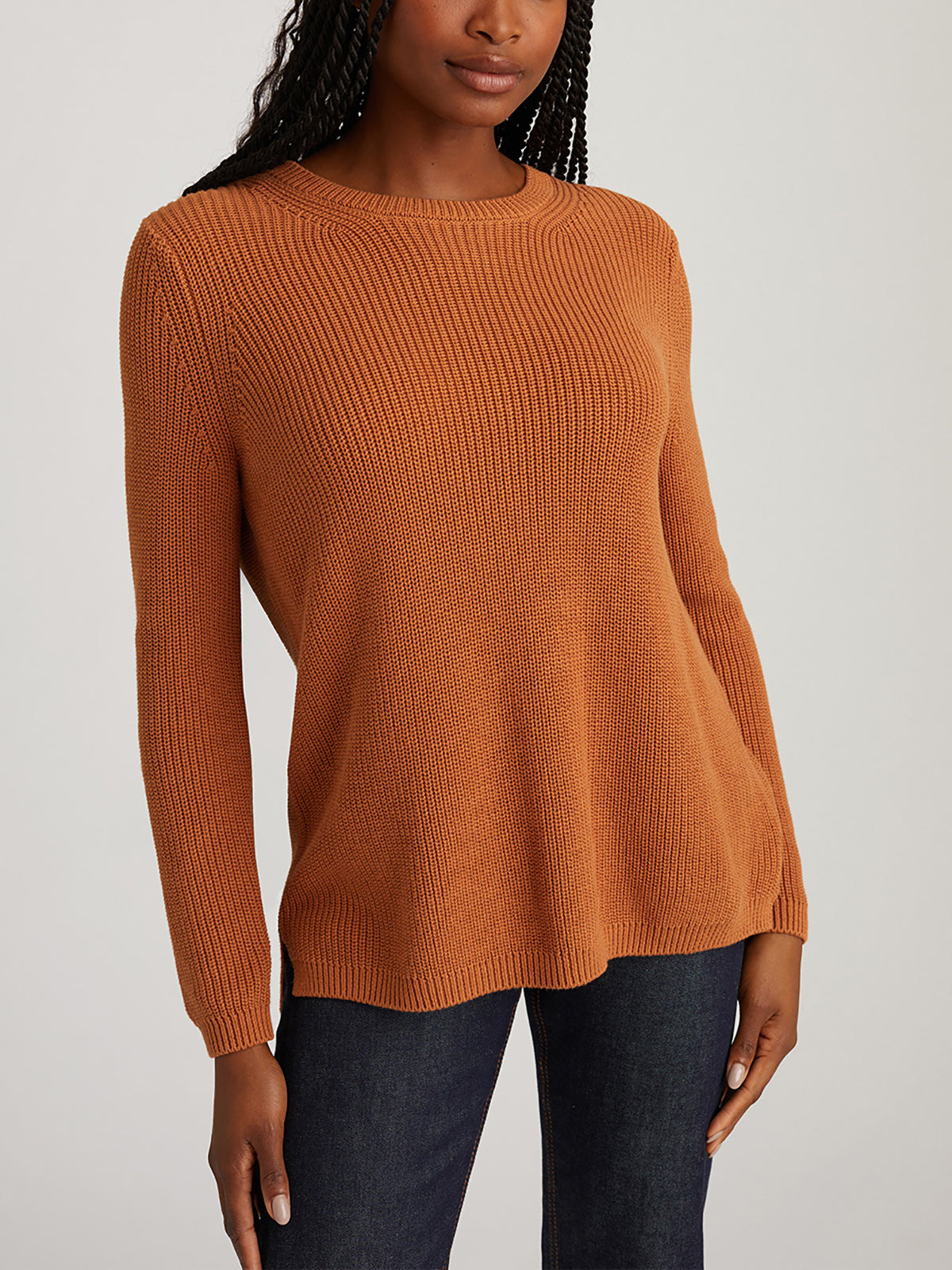 A person wears a rust-colored ribbed sweater with a shaker stitch from 525 America and dark jeans against a neutral backdrop. The versatile, relaxed-fit sweater suits any occasion. Their long hair is styled in braids.