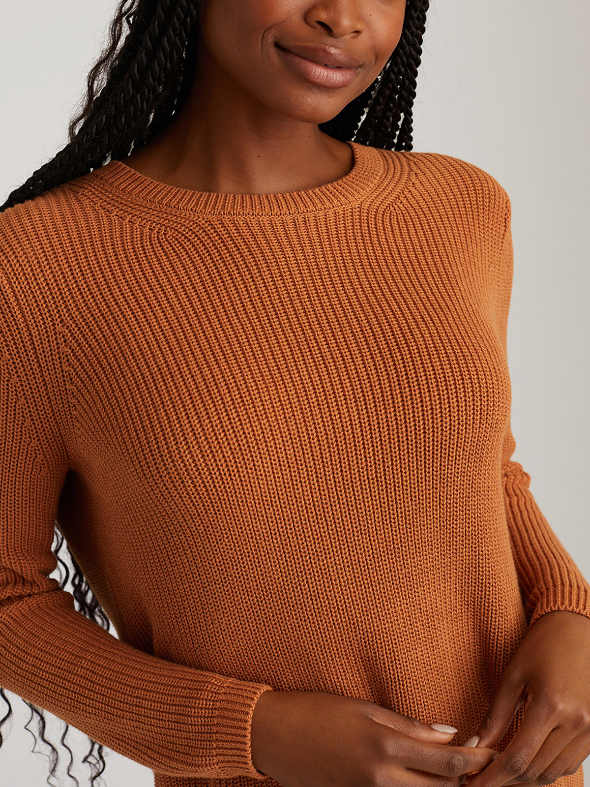 A person with long braided hair wears a rust-colored, rib-knit sweater from 525 America in a relaxed fit. They pose against a neutral background with arms slightly bent, hands on waist, and a partial smile, highlighting their versatile style.