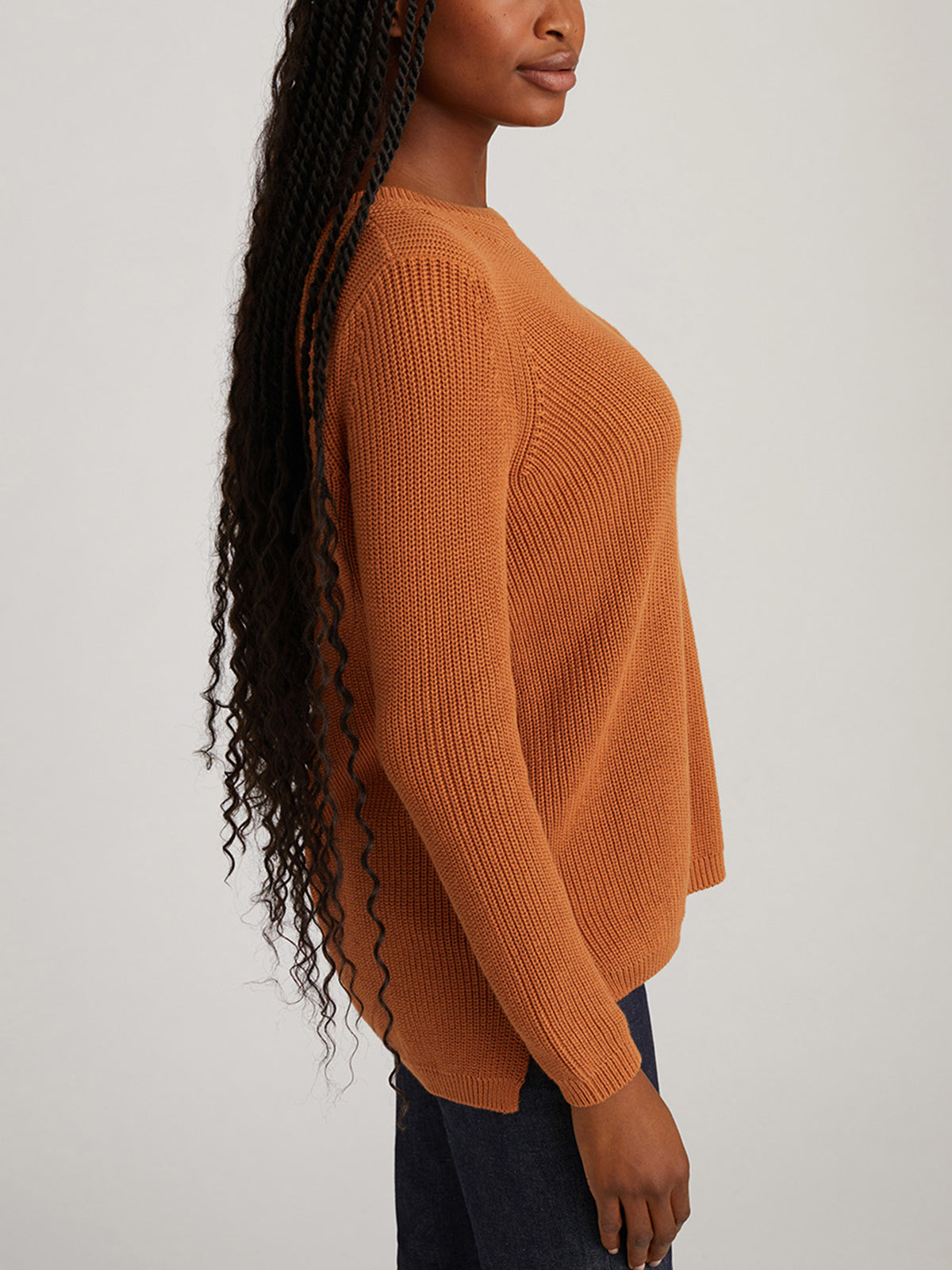 A person with long, wavy hair is wearing a relaxed-fit ribbed sweater in rust by 525 America paired with dark pants. They stand sideways against a plain background, highlighting their versatile style.
