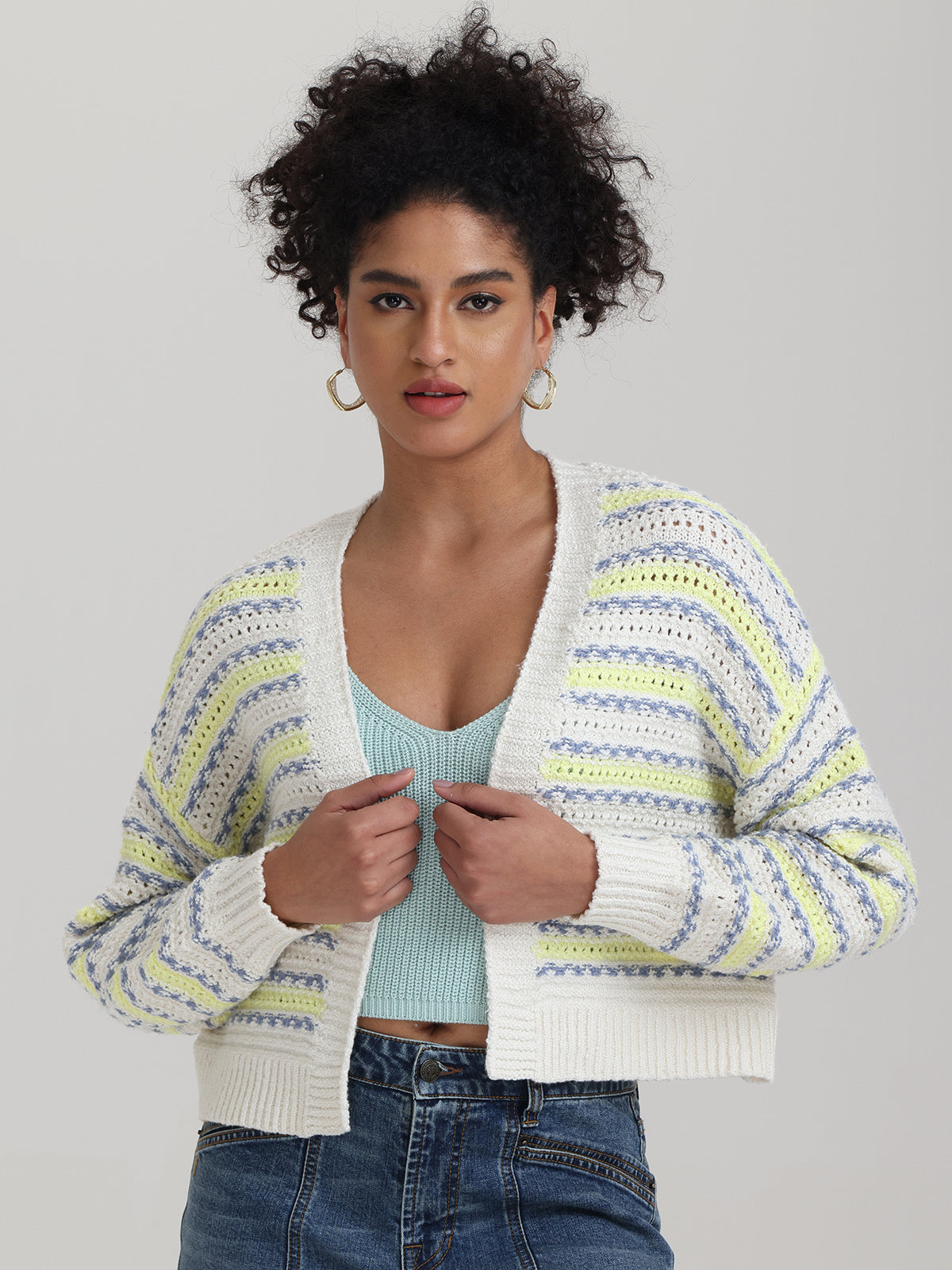 A person with curly hair wears a striped 525 America cardigan over a light blue top and jeans. The open design features eye-catching white, blue, and yellow stripes against a plain, light-colored background.