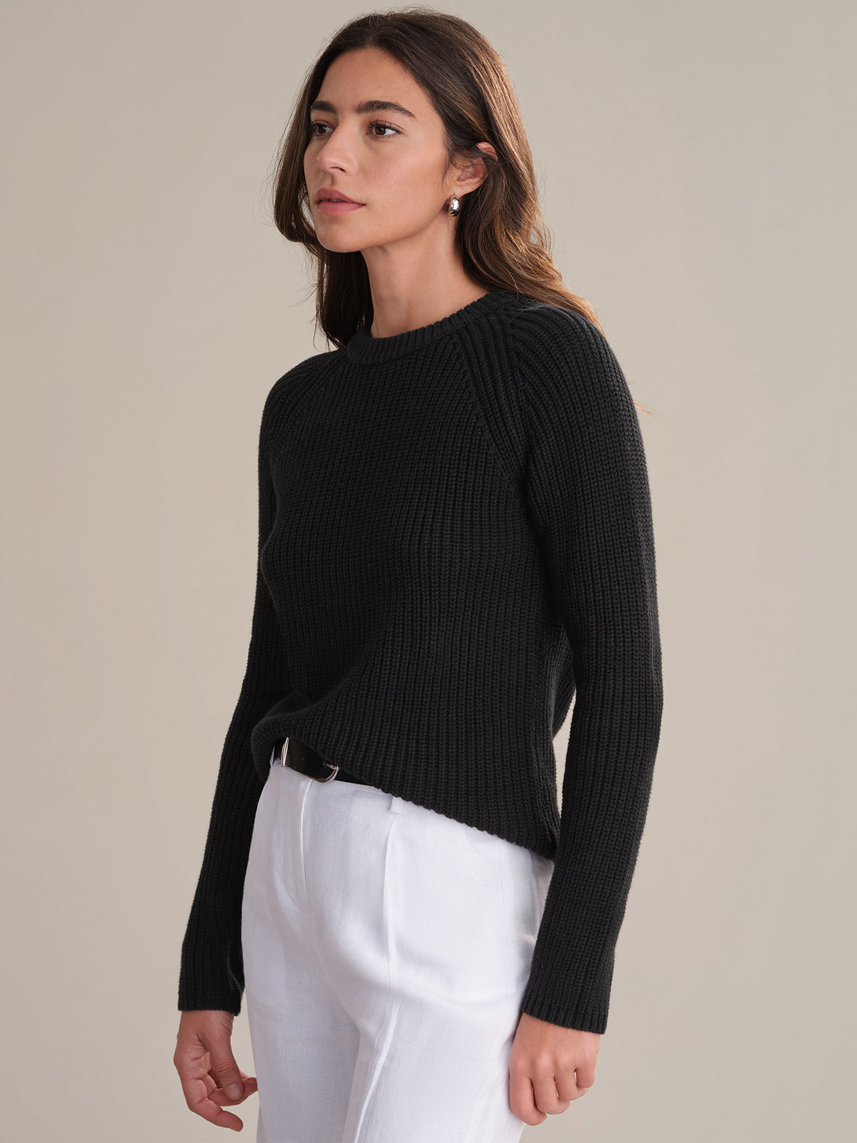 A woman with long brown hair wears a black ribbed knit raglan sleeve sweater by 525 America and white pants, standing against a plain beige background and gazing slightly to the side with a neutral expression.