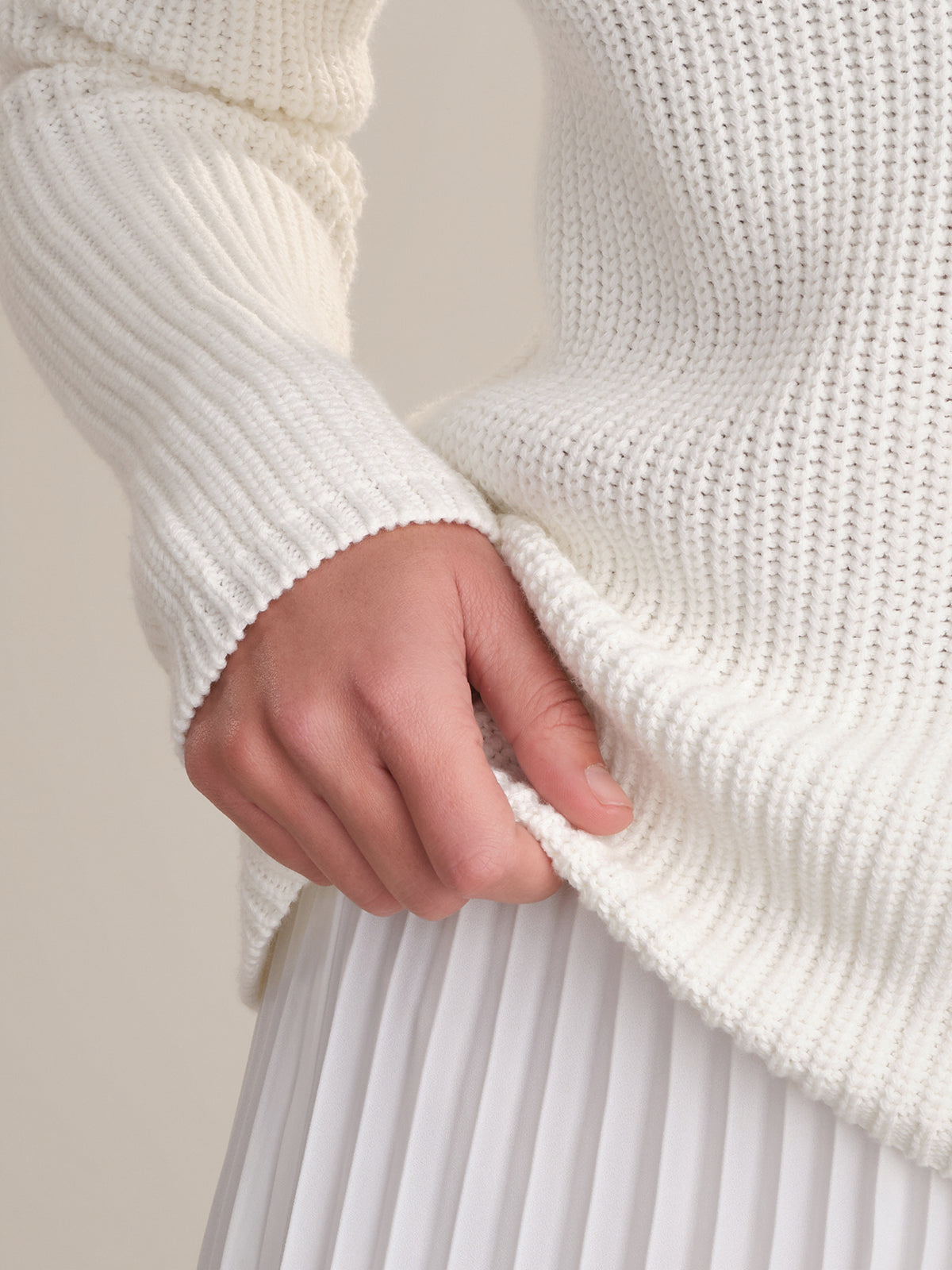 A close-up of a person wearing a 525 America white cotton shaker stitch sweater with raglan sleeves and a white pleated skirt, holding the sweater’s hem against a plain, neutral background.