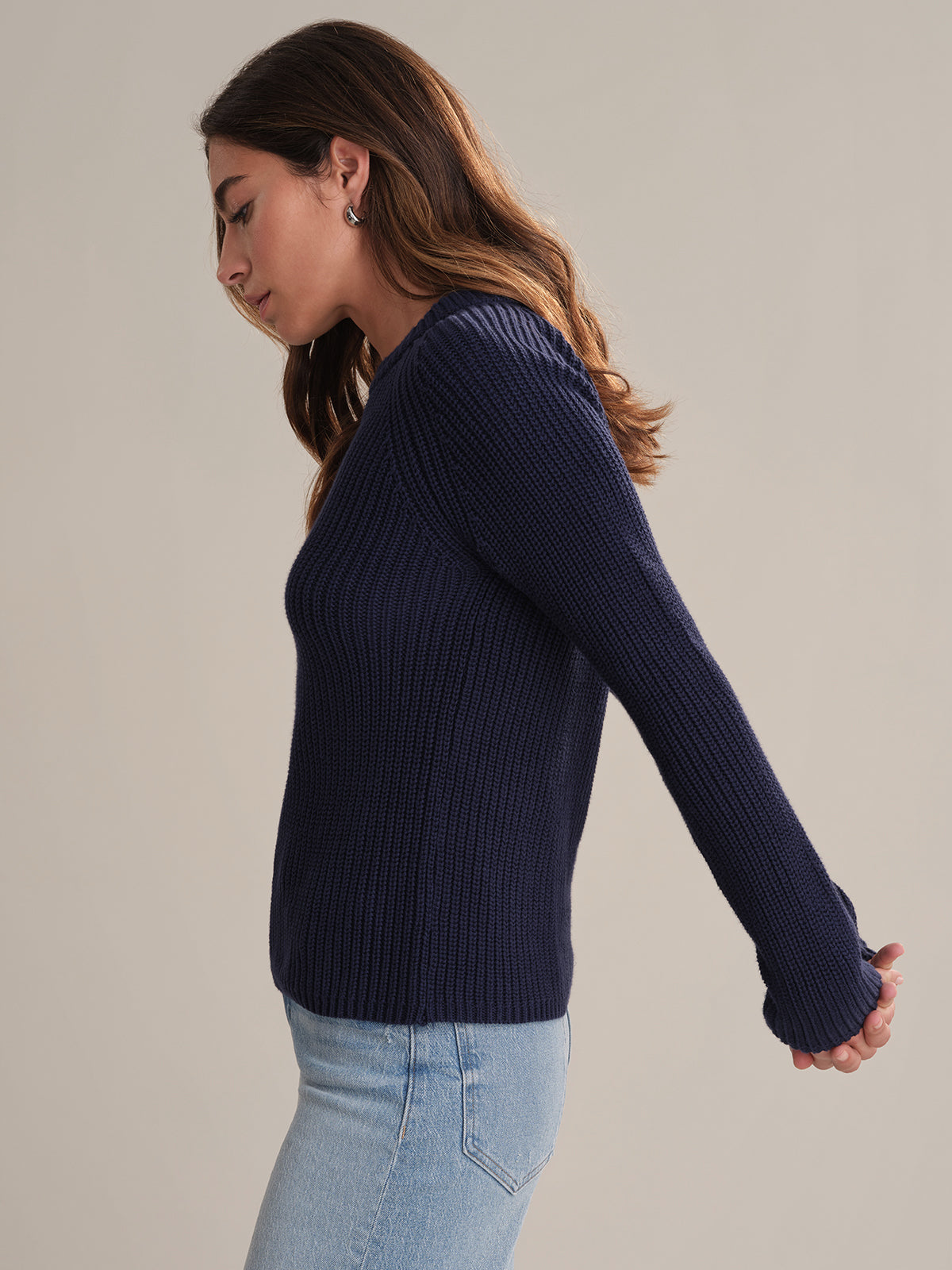 A woman with long brown hair models a navy blue 525 America cotton shaker stitch sweater with a fitted silhouette and light blue jeans. She stands sideways, arms extended behind her, against a plain light background.