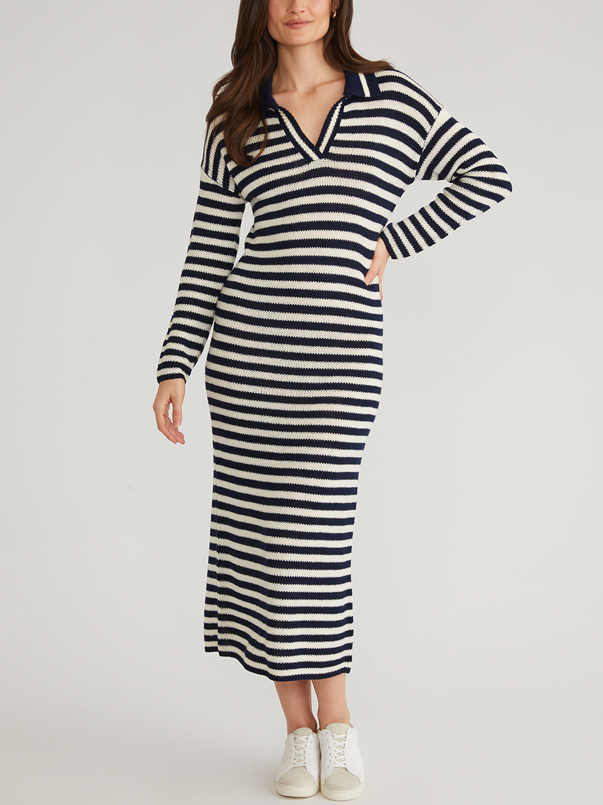 A woman poses in a navy and white striped knit dress by 525 America, featuring long sleeves, a v-neckline, and collar. Styled with white sneakers against a plain backdrop, its an Instagram-favorite ideal for transitional weather.