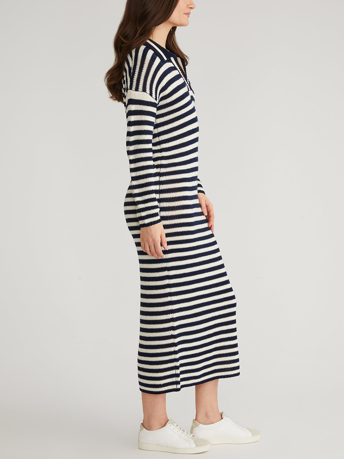 A woman wearing a navy and white sailor-stripe motif, long-sleeve knit dress by 525 America stands sideways, paired with white sneakers. Her long brown hair cascades down as she poses against a plain, light-colored background. This Instagram viral dress is ideal for transitional weather.