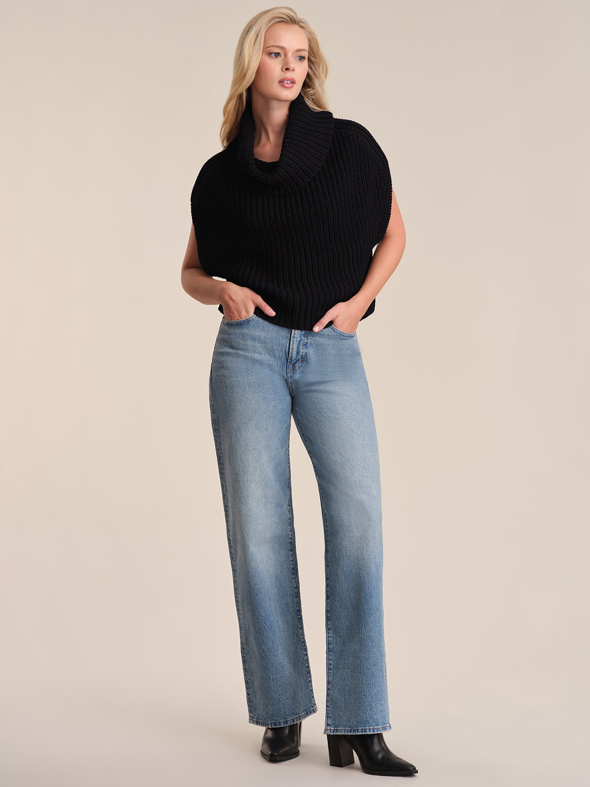 A person stands against a beige background wearing a 525 America sleeveless black turtleneck chunky neck sweater, light blue relaxed fit straight-leg jeans, and black heeled boots, hands in pockets with a neutral expression.