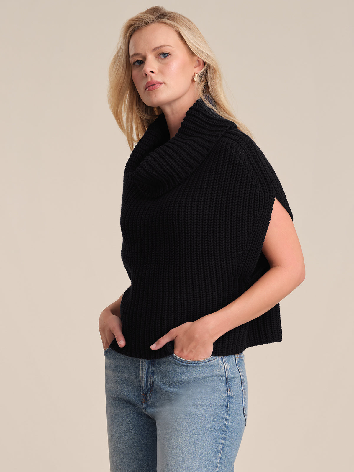 A person with long blonde hair models a stylish black chunky neck sweater from 525 America and light blue jeans, standing against a plain beige background with hands in their pockets.