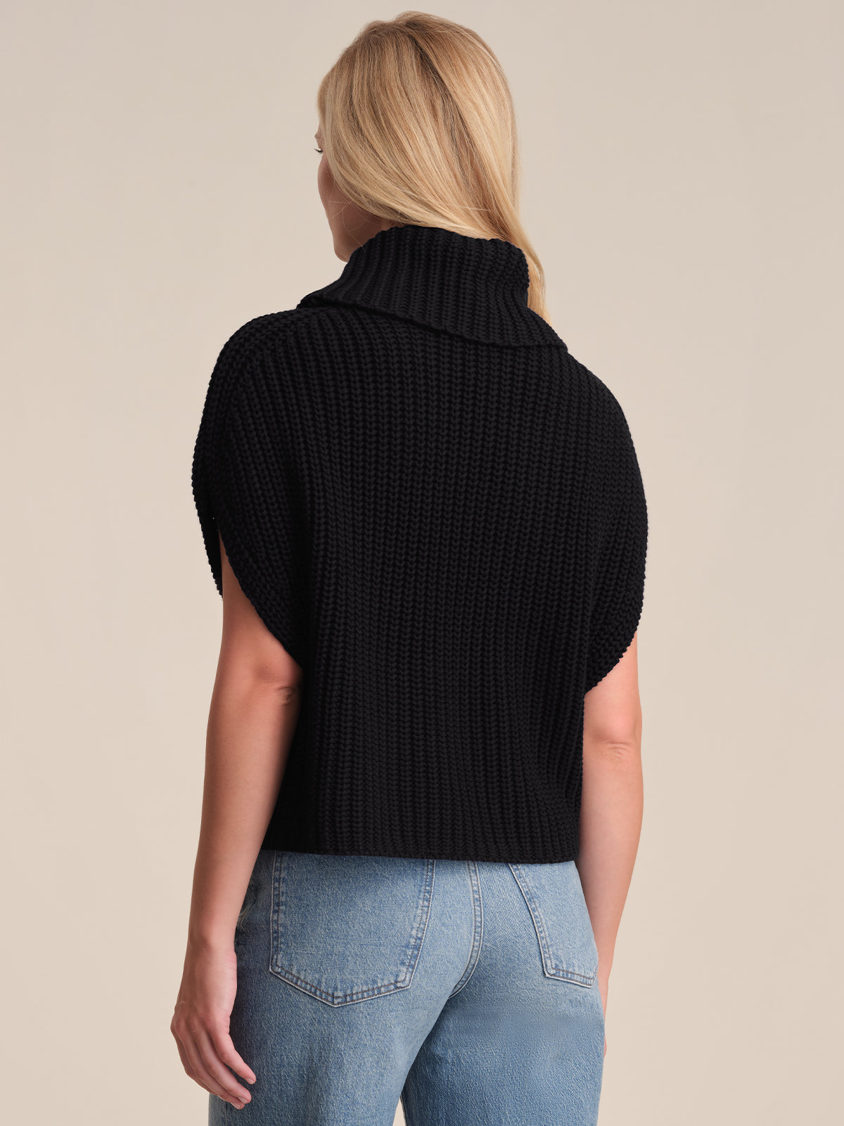 A person with long blonde hair is seen from behind, modeling a 525 America relaxed fit black knit short-sleeve turtleneck sweater and blue high-waisted jeans against a plain light background.