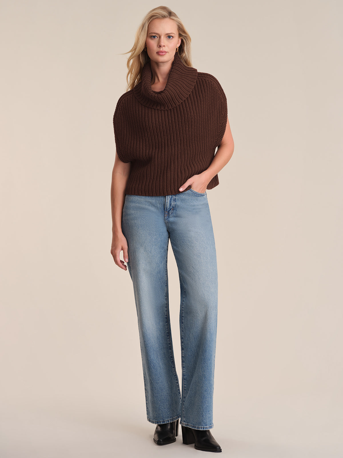 A woman stands facing forward in a chic look, wearing a 525 America dark brown chunky neck cowl sweater, light blue high-waisted relaxed-fit jeans, and black boots against a beige background. One hand rests in her pocket.