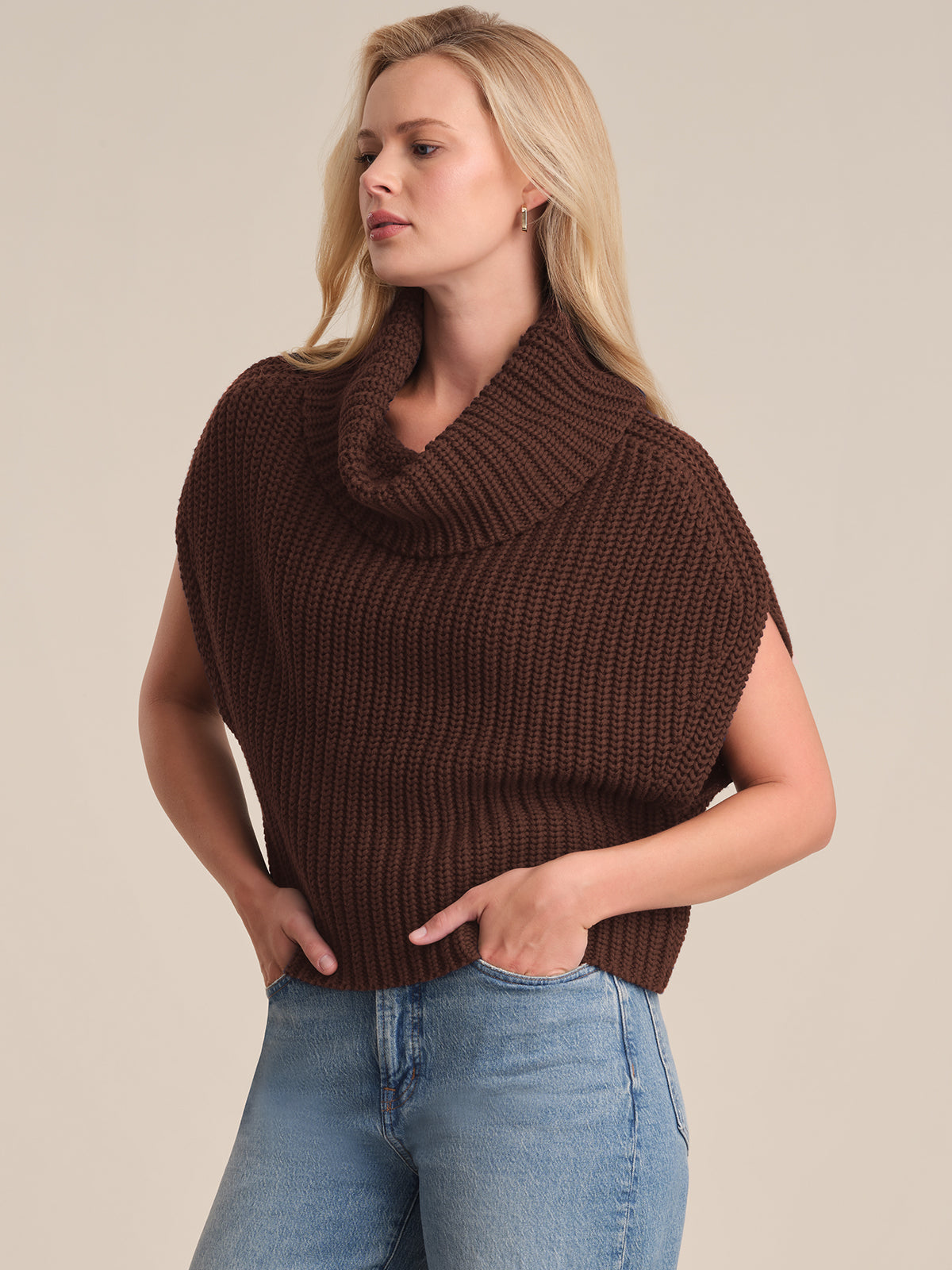 A person with long blonde hair wears a 525 America sleeveless chunky brown knit sweater featuring a wide cowl neck and relaxed fit, paired with light blue jeans, standing against a plain beige background.