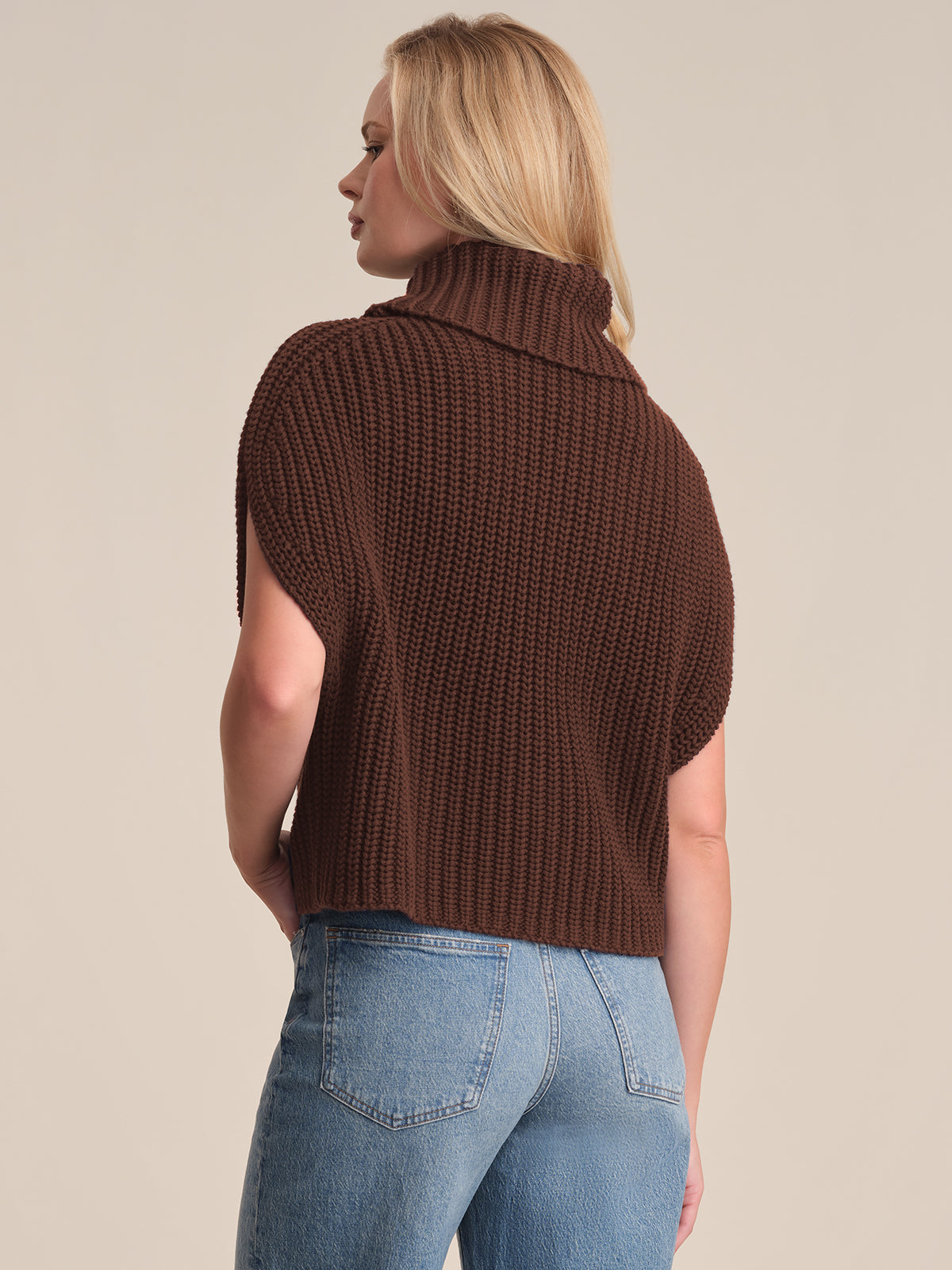 A person with blonde hair stands sideways, back to the camera, against a beige background, wearing a 525 America relaxed fit chunky brown short-sleeved turtleneck sweater and blue jeans.