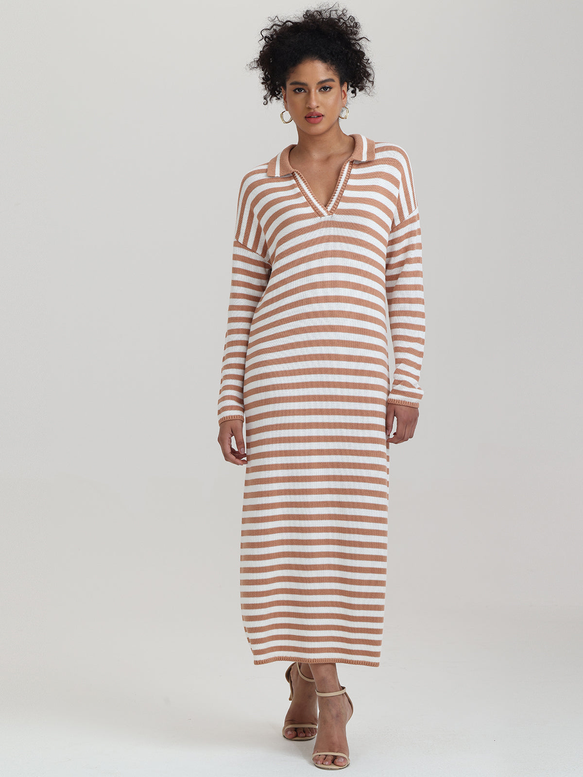 Wearing a relaxed-fit, long-sleeved knit dress by 525 America with tan and white sailor stripes, a person enjoys transitional weather. The dress features a V-neckline and collar. Their curly hair complements beige high-heeled sandals against a plain background.