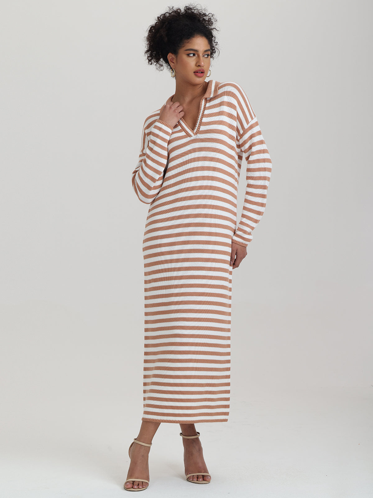 A person wears a 525 America long, brown and white striped dress with a relaxed fit, long sleeves, and a V-neck, paired with beige high-heeled sandals. Ideal for transitional weather, the background is plain and light-colored.
