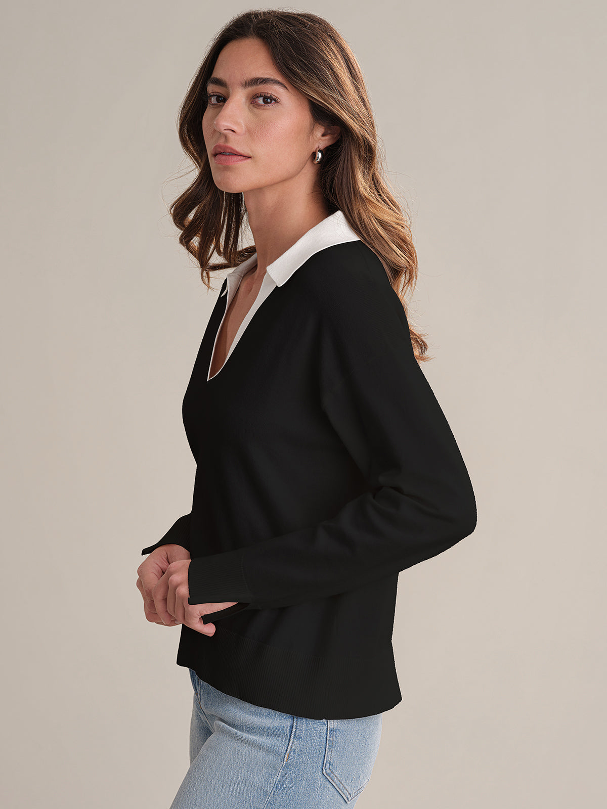 A person with long brown hair wears a black 525 America lightweight pullover with a white Johnny collar and light blue jeans, standing sideways and looking at the camera against a plain background.