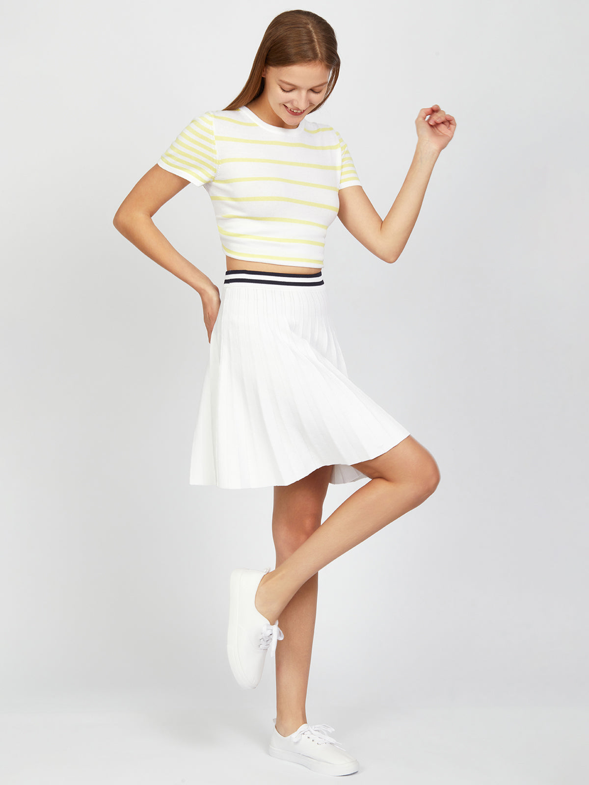 In a stylish blend of sporty-chic, this person dons a yellow and white striped short-sleeve top by 525 America with a white pleated skirt and sneakers. Gracefully posed against a plain backdrop, they balance on one leg, slightly bending the other, while their relaxed smile enhances the stripe-waist look.