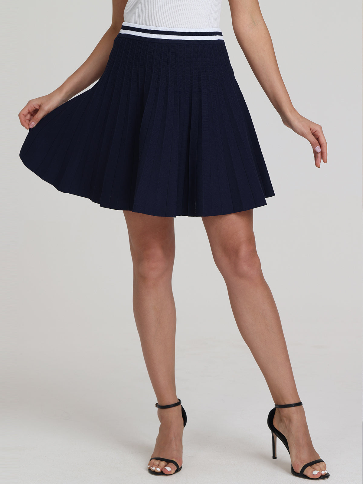 A person wears a 525 America white sleeveless top and a navy pleated skirt with a striped waist, paired with black high-heeled sandals. They strike a pose by holding part of the skirt, adding sporty-chic flair.