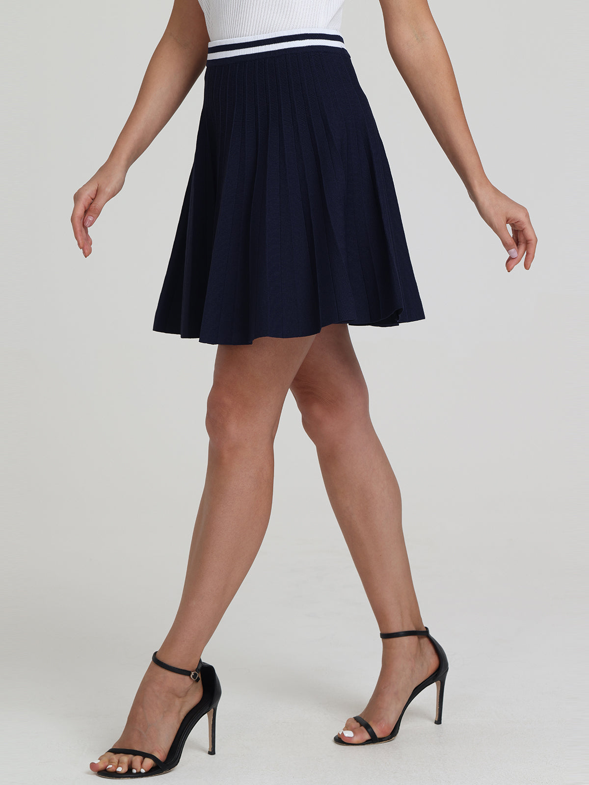 A person in a pleated, short dark blue skirt from 525 America and black high-heeled sandals embodies a sporty-chic vibe against a plain white background.