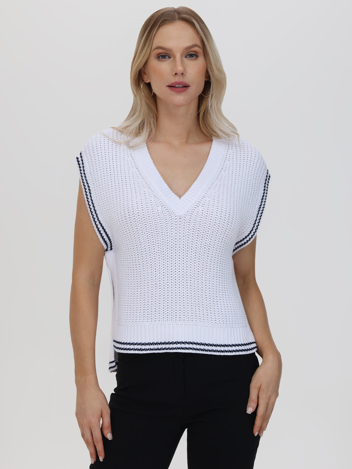 A person with long hair wears a sophisticated white knit top from 525 America, showcasing a cable-knit design and black trim on the neckline and sleeves. The deep V-neck style pairs elegantly with black pants against a plain, light-colored backdrop.