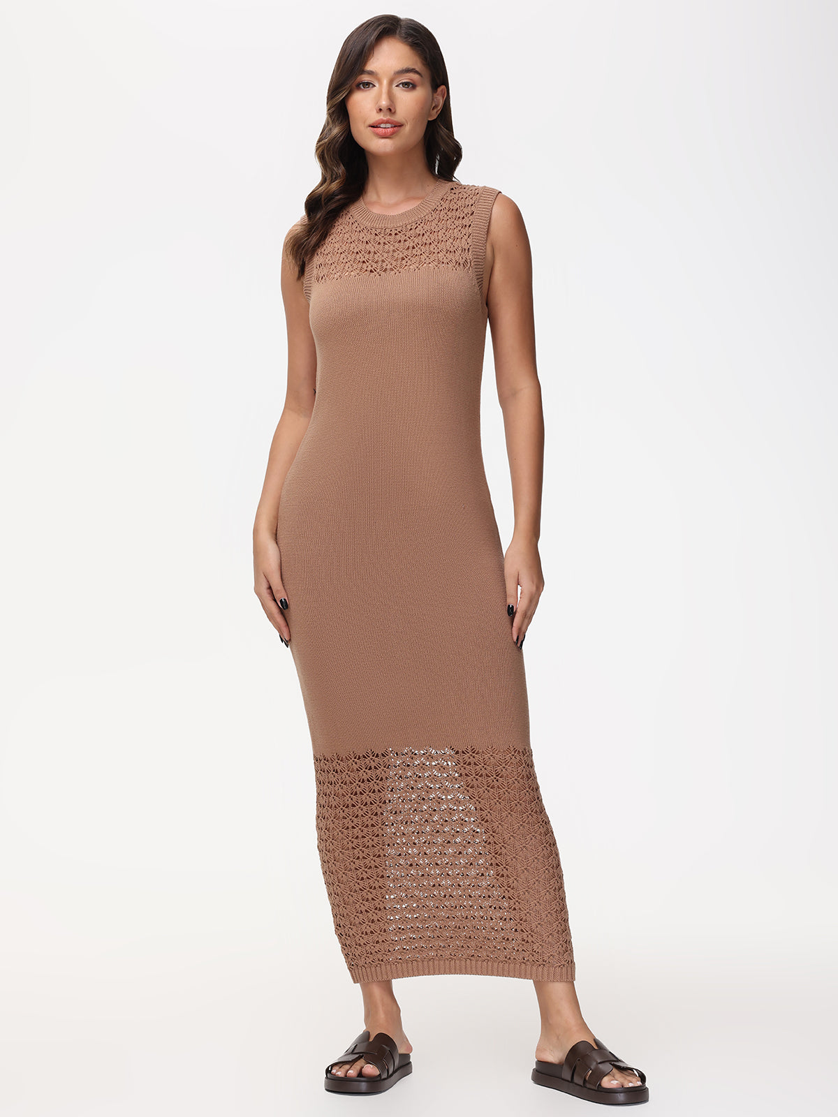 Adorned in a sleeveless, brown crochet maxi dress by 525 America, a woman embodies bohemian charm against a plain white background. Featuring intricate patterns at the top and bottom, she completes her look with black sandals and loose waves—ideal for summer soirees.