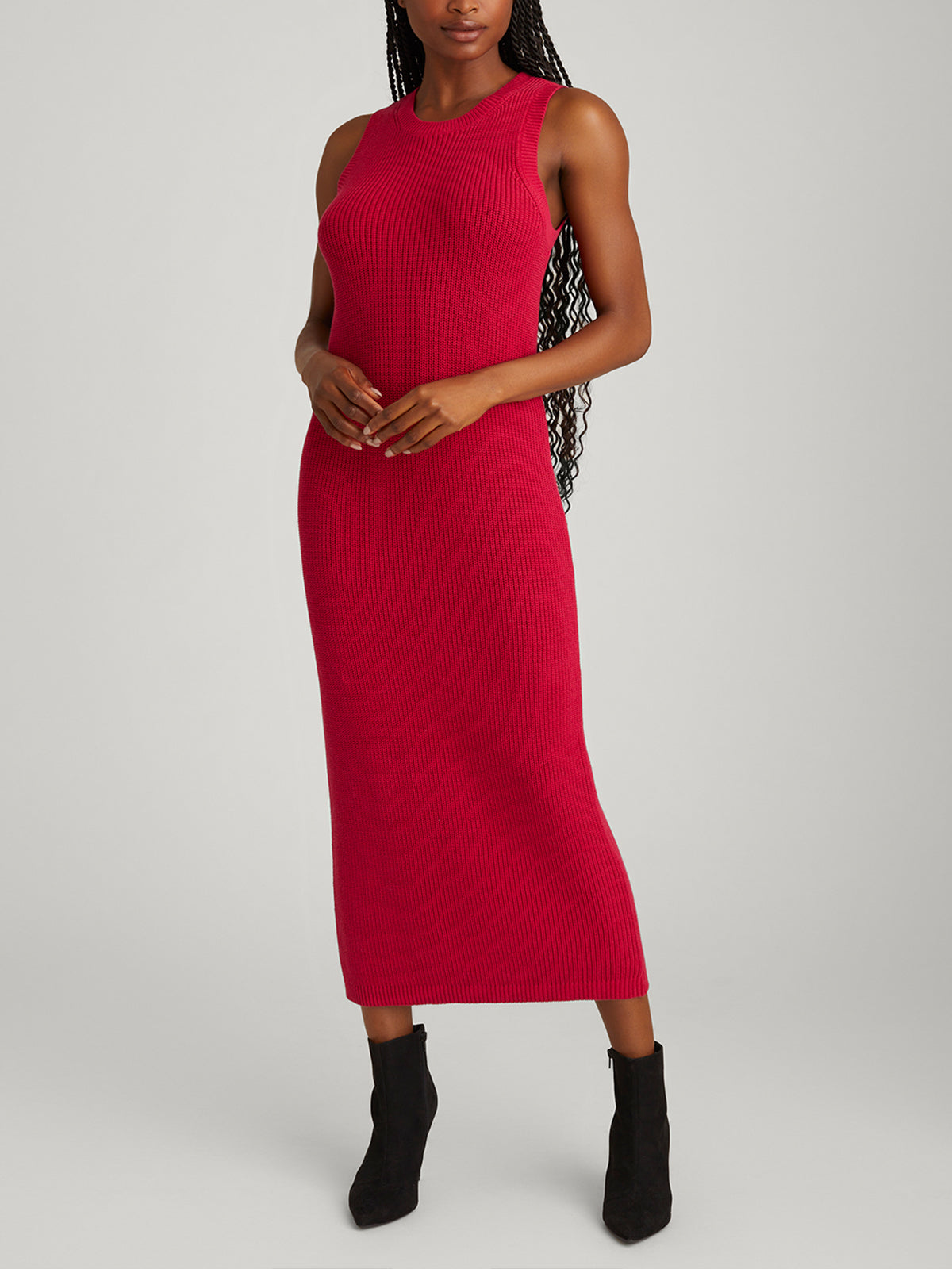 Wearing the 525 Americas Emma Crewneck Shaker Stitch Tank Dress, a stylish red sleeveless knit made of 100% cotton, someone stands against a gray background. The dresss ribbed texture pairs well with their long braided hair and black ankle boots.