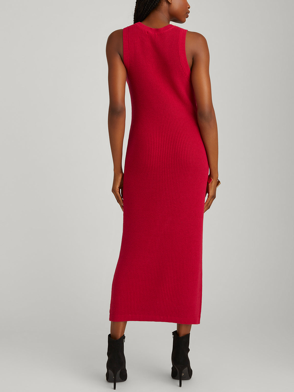 The image shows a person with their back to the camera in a chic ensemble. Theyre in the 525 America Emma Crewneck Shaker Stitch Tank Dress, vivid red, made from 100% cotton. Black ankle boots complement the look against a plain gray background.