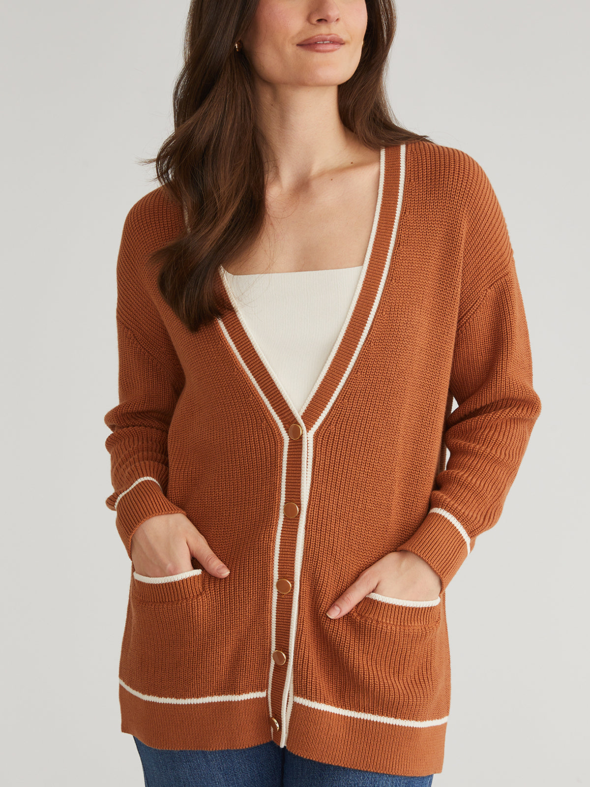 A person wears a relaxed-fit, rust-colored 525 America grandpa-style cardigan with white trim and pockets over a white top. The oversized, buttoned cardigan has hands-in-pockets, against a plain light-colored background enhancing the cozy look.