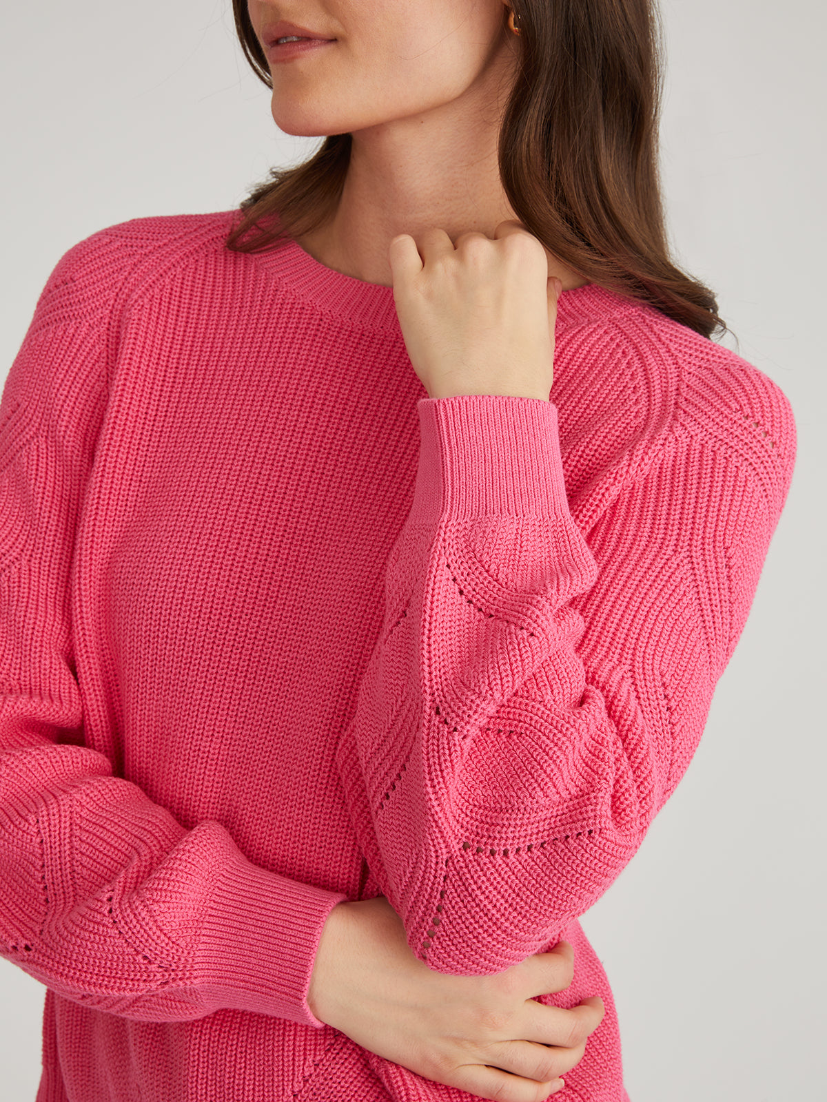 A person with long brown hair wears a bright pink 525 America knit sweater with raglan pointelle sleeves. This soft, 100% cotton fabric features a textured pattern as they hold an arm near their chin against a plain, light background.