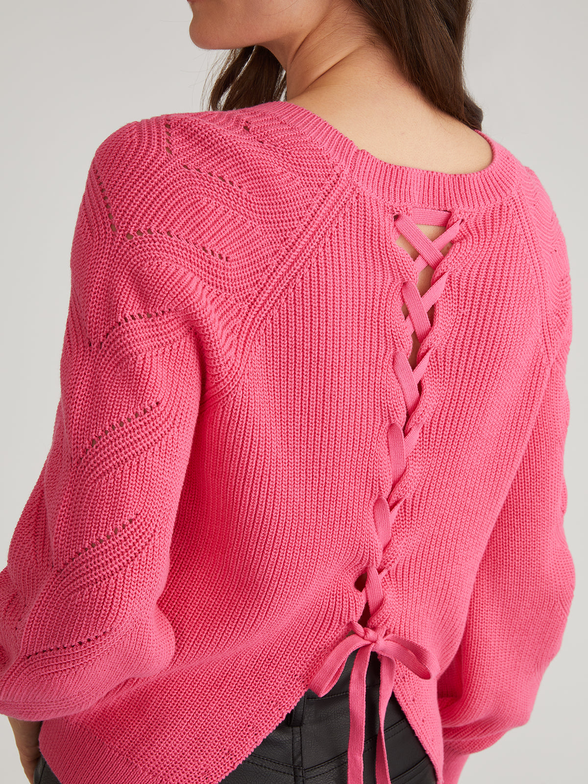 A woman with long brown hair stands against a plain background wearing 525 Americas bright pink Lace Back Pullover. Made from 100% cotton, this sweater features a lace-up back, hole patterns, ties at the bottom, and Raglan Pointelle Sleeves.