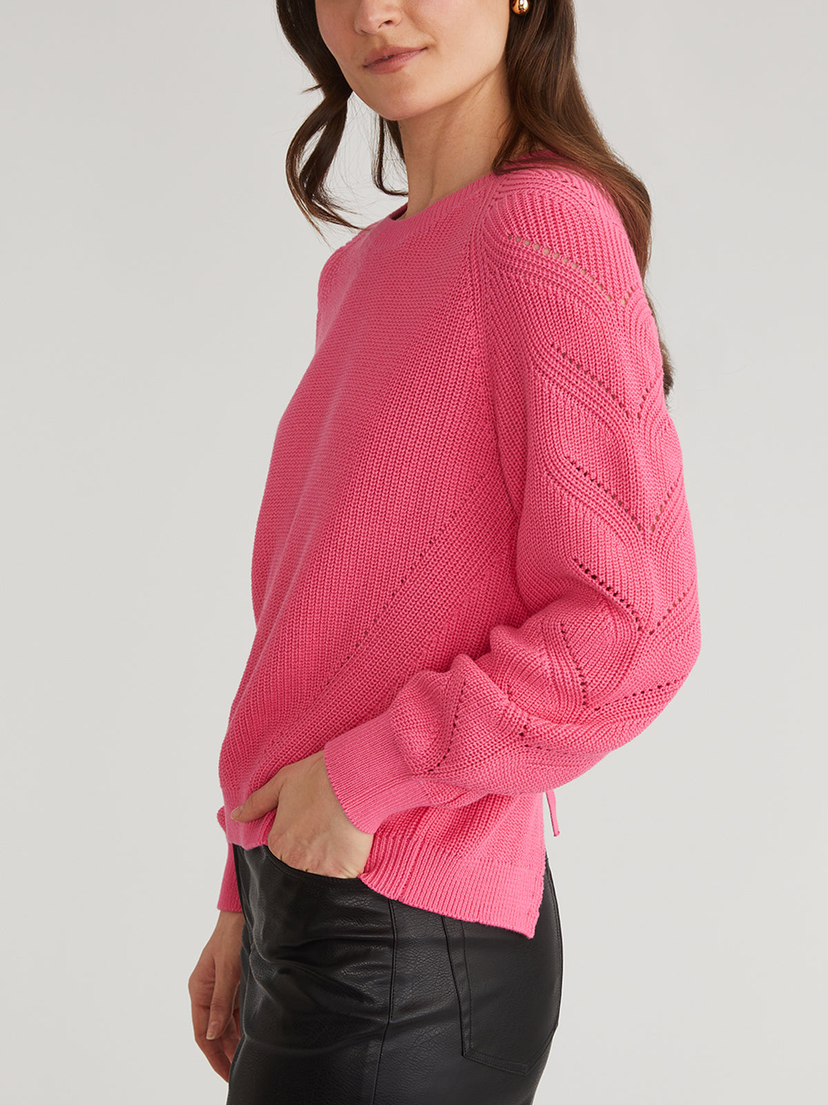A woman in a 525 America bright pink knit sweater with raglan pointelle sleeves and black leather pants stands in profile, her left hand in her pocket. The 100% cotton sweaters textured pattern adds flair against the plain, light-colored background.