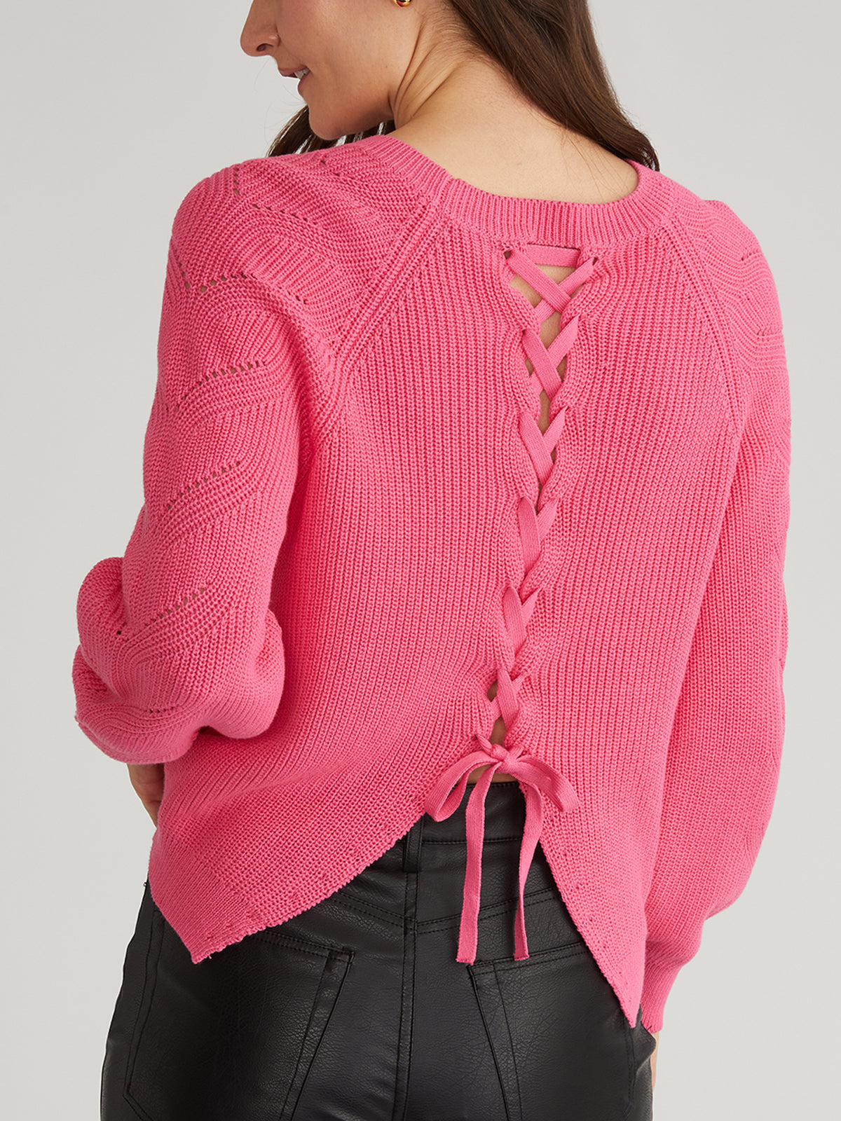 The person wears a pink Lace Back Pullover by 525 America, featuring an intricate lace-up design with crisscross tied ribbons. This 100% cotton sweater has a slightly uneven hem, paired stylishly with black leather pants, highlighting the detailed back view.