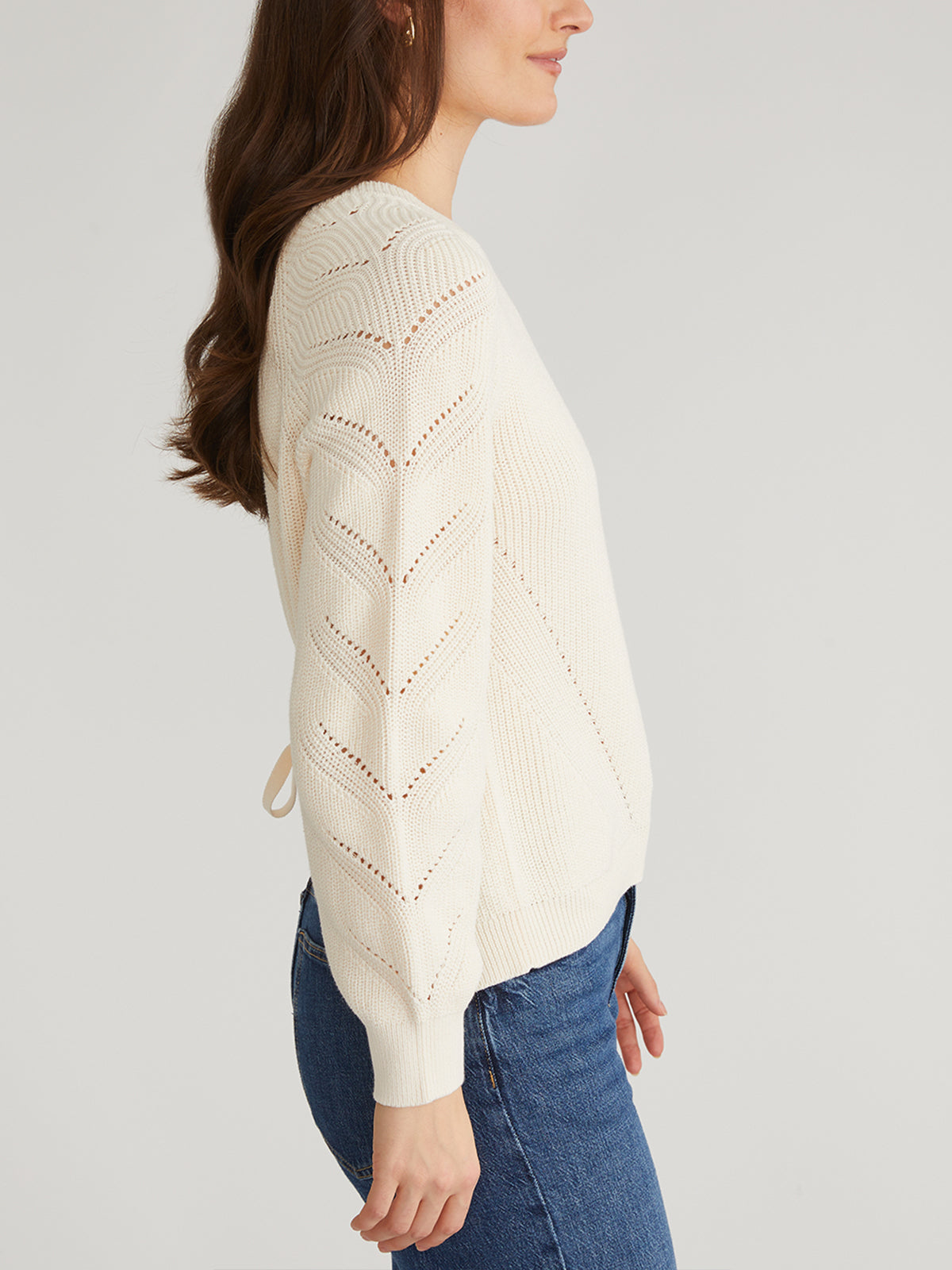 A person with long brown hair wears a cream-colored 525 America sweater featuring Raglan Pointelle Sleeves, paired with blue jeans, and stands in profile against a plain light gray background.