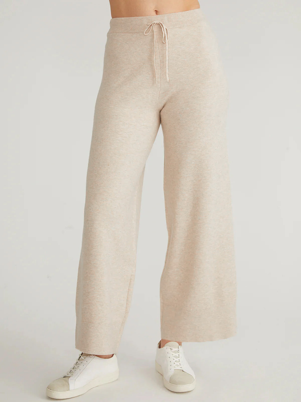 Wearing 525 Americas beige high-rise wide-leg pants and white sneakers, a person stands against a plain backdrop. The focus is on the lower body, highlighting the silhouettes relaxed elegance and emphasizing the pants comfortable, casual style.